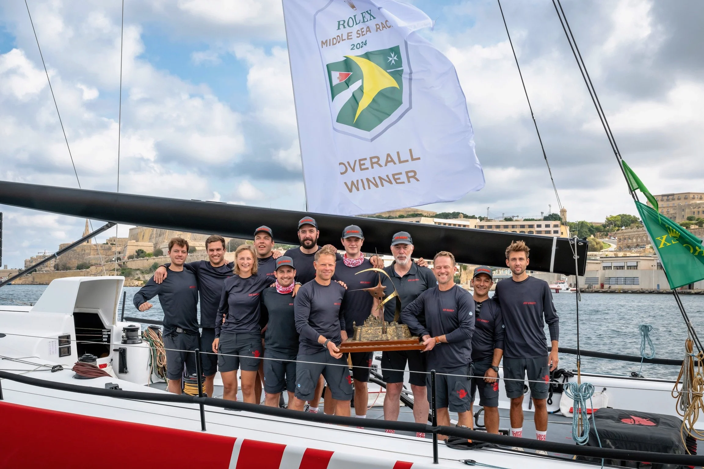 A group of ten people on a sailboat holding a trophy, celebrating their overall win in a race. A large flag with the Rolex Middle Sea Race 2024 logo and 'Overall Winner' text is displayed overhead. The background shows a cityscape and cloudy sky.