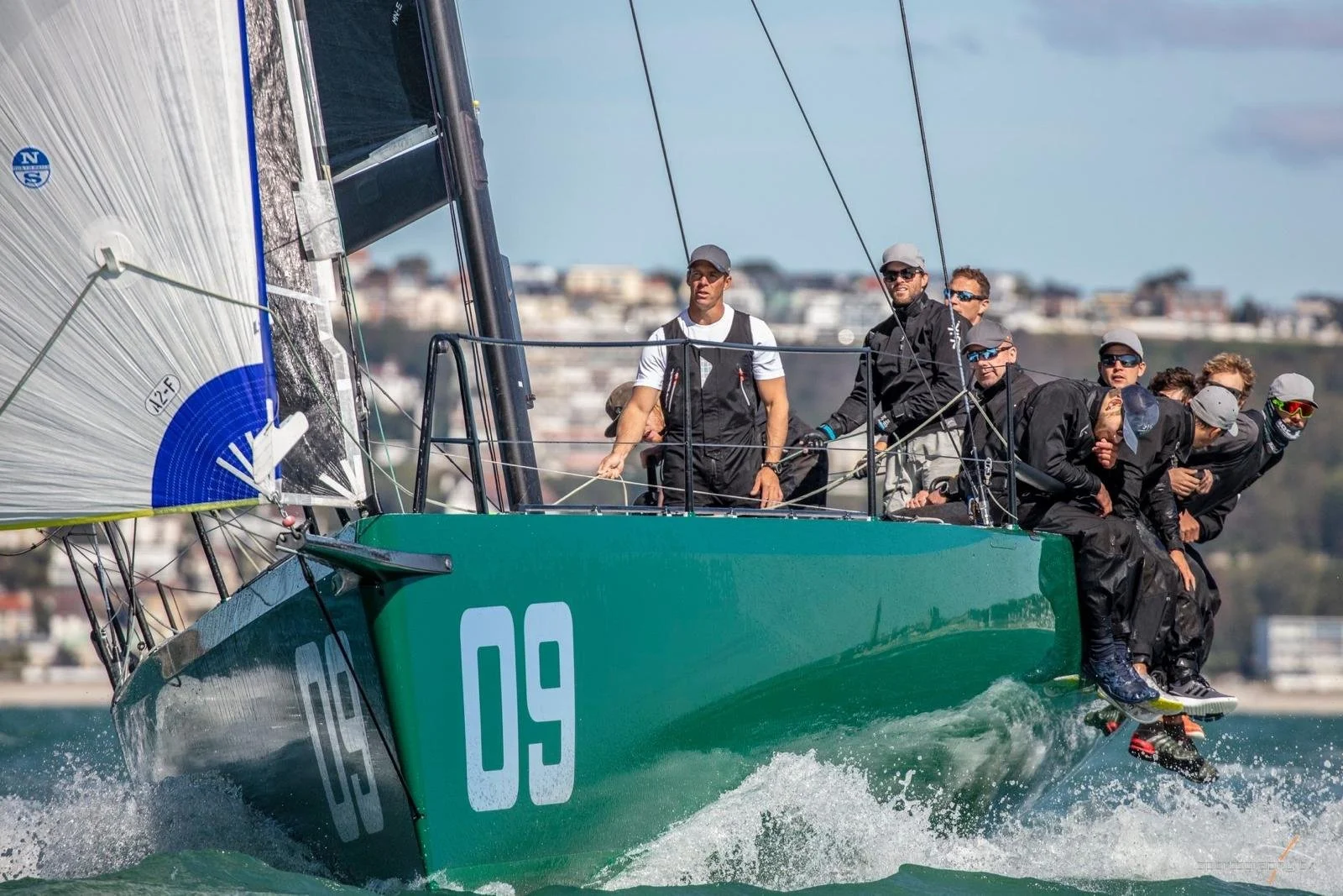 A group of sailors racing on a green sailboat with the number 09, leaning over the side for stability in a regatta on the water, with a cityscape in the background.