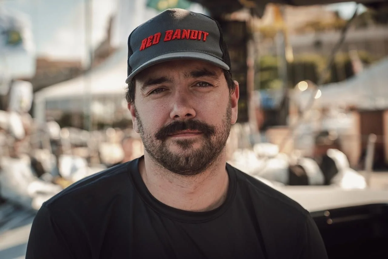 A man with a beard and mustache wearing a gray baseball cap with red embroidery that says 'RED BANDIT' and a black t-shirt outdoor during daytime with a blurred background of boats and water.