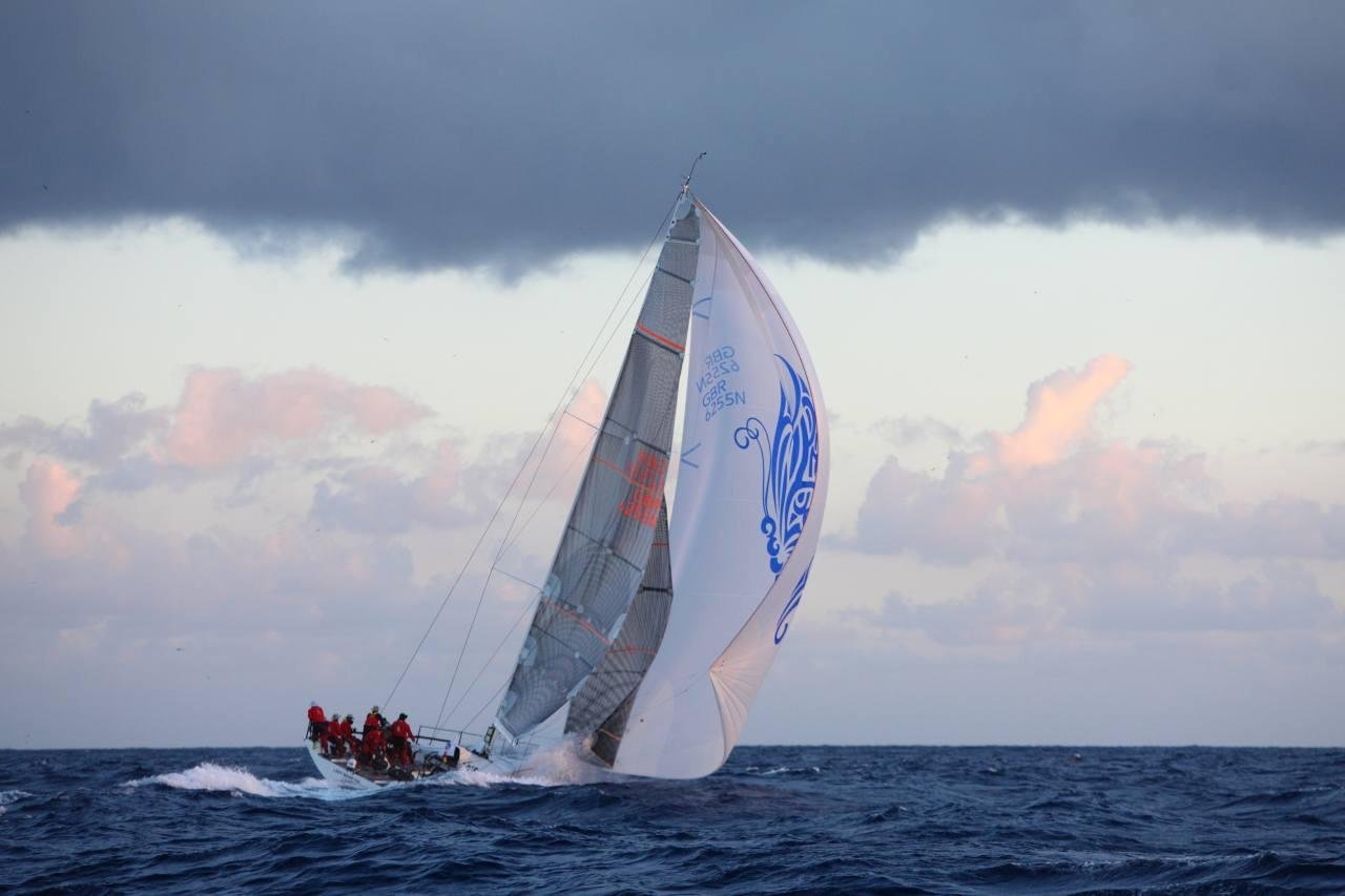 A sailboat with large sails, including one with a blue design, racing on the ocean with dark clouds overhead.