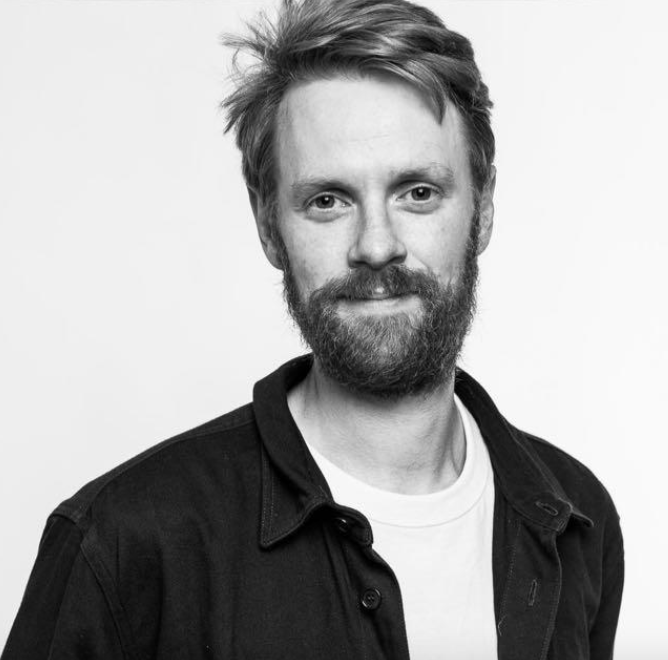 Schwarz-Weiß-Fotografie eines Mannes mit Bart und ungekämmtem Haar, trägt ein dunkles Hemd und ein weißes T-Shirt, vor hellem Hintergrund.