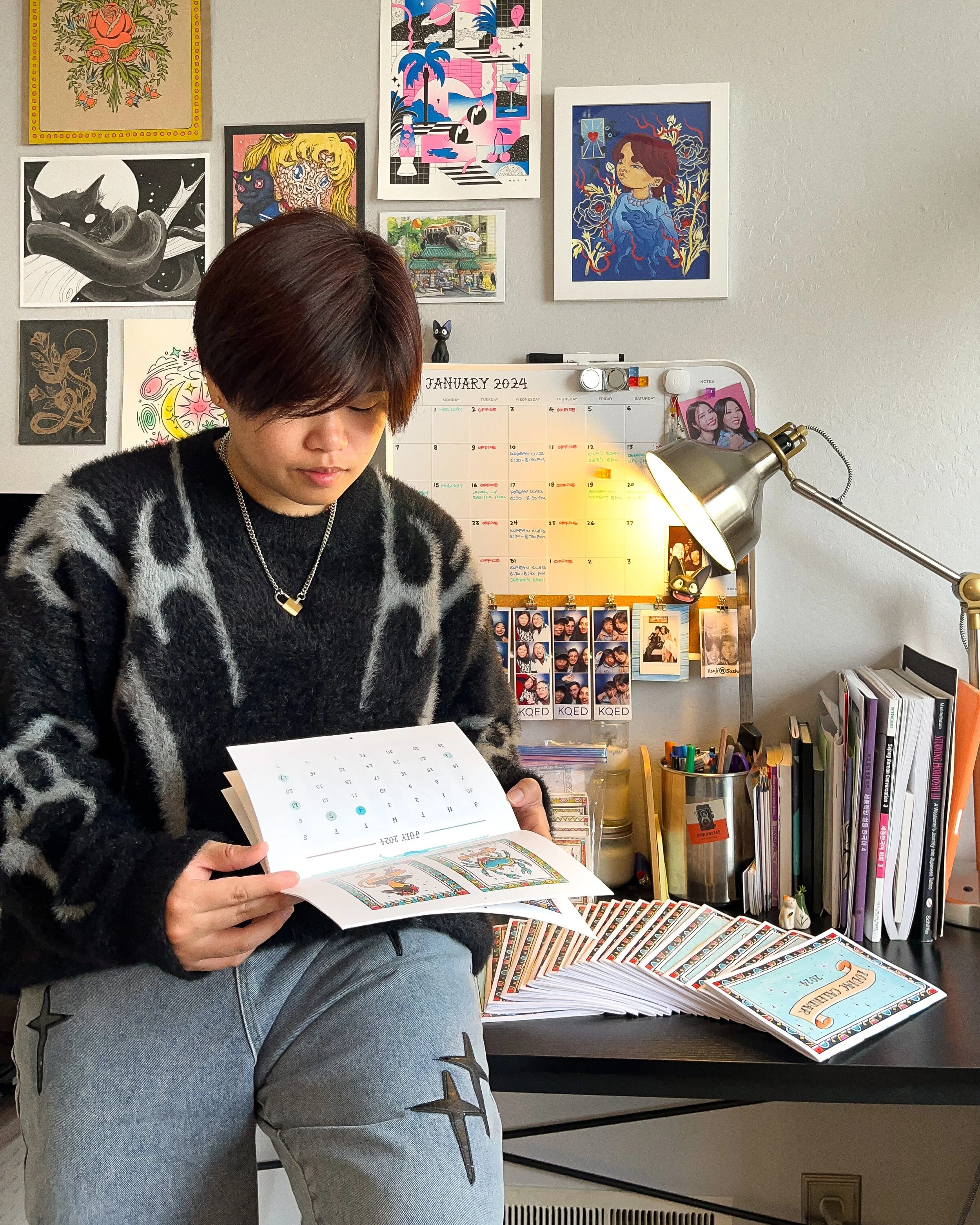 An Asian-American person with short, dark brown hair looks down at a calendar in their hands with a soft, subtle smile. They are wearing a black sweater, blue jeans and a silver chain necklace. The wall behind them is covered in art.
