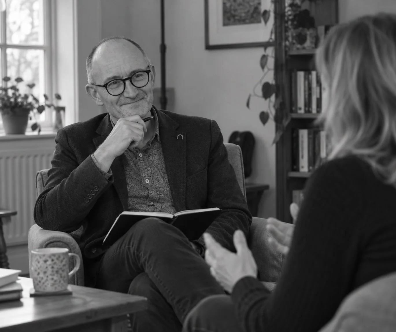 An older man with glasses and a beard, wearing a blazer, is sitting in a comfortable chair, holding a notebook and a pen. He is listening attentively to a woman with long hair, who is talking to him with her hands. The setting appears to be a cozy room, possibly an office or a home, with bookshelves and pictures on the wall.