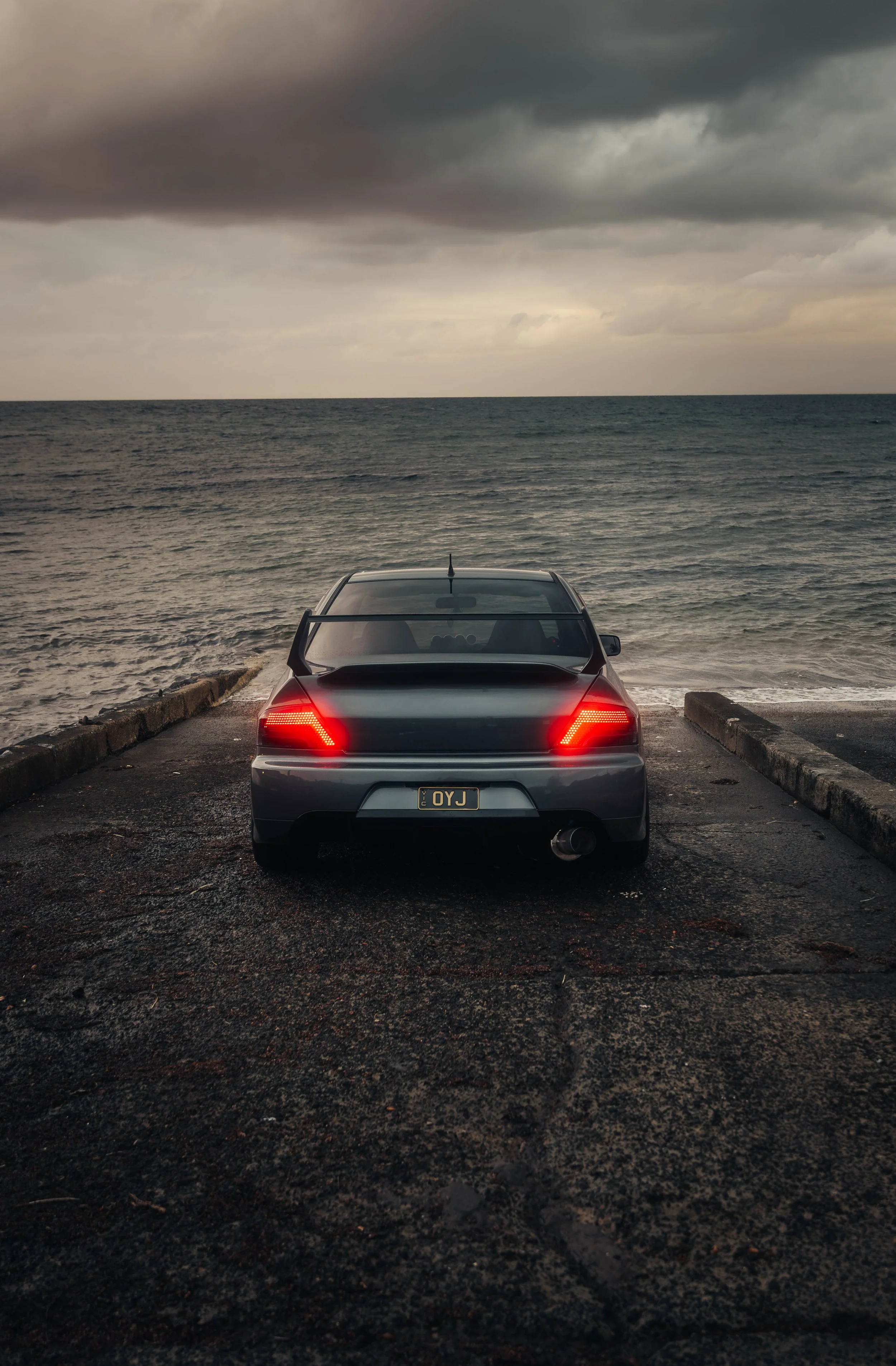 A dark-colored sports car with red taillights parked at a seaside dock, facing the ocean under a cloudy sky. EVO 8, evo 9 Mitshubishi evolution, jdm car, jdm photo, work wheels, work racing, race car, speed, jdm. 