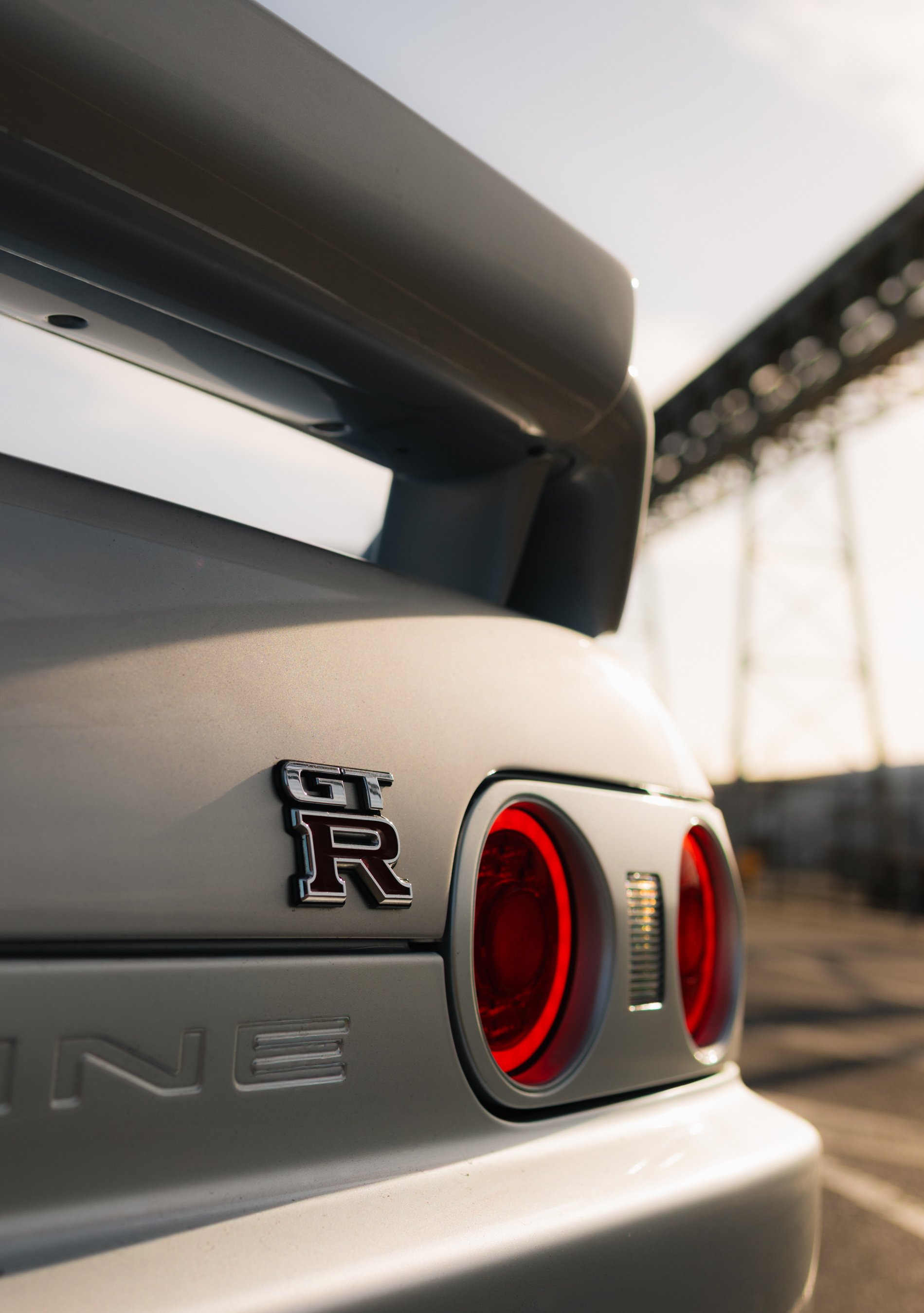 Close-up of the rear of a light gray Nissan GT-R sports car, showing the GT-R badge and round red taillights, with a blurred industrial structure in the background. Nissan R32 GTR Skyline, GTR photo, dock photo, car photos, jdm at the docks, melbourn