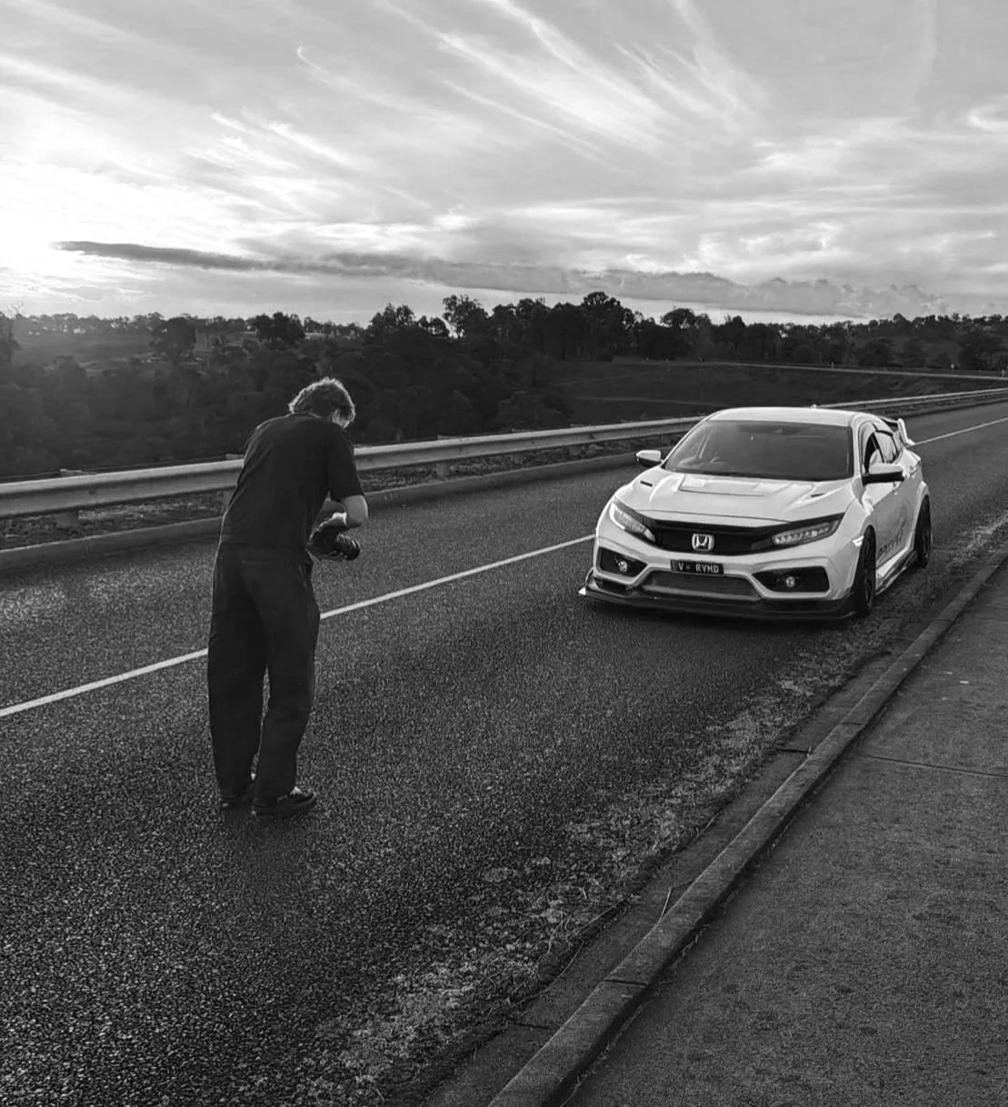 A photographer is taking photos of a modern Honda fk8 type r civic parked on the side of a highway during sunset with a cloudy sky and scenic landscape in the background.
