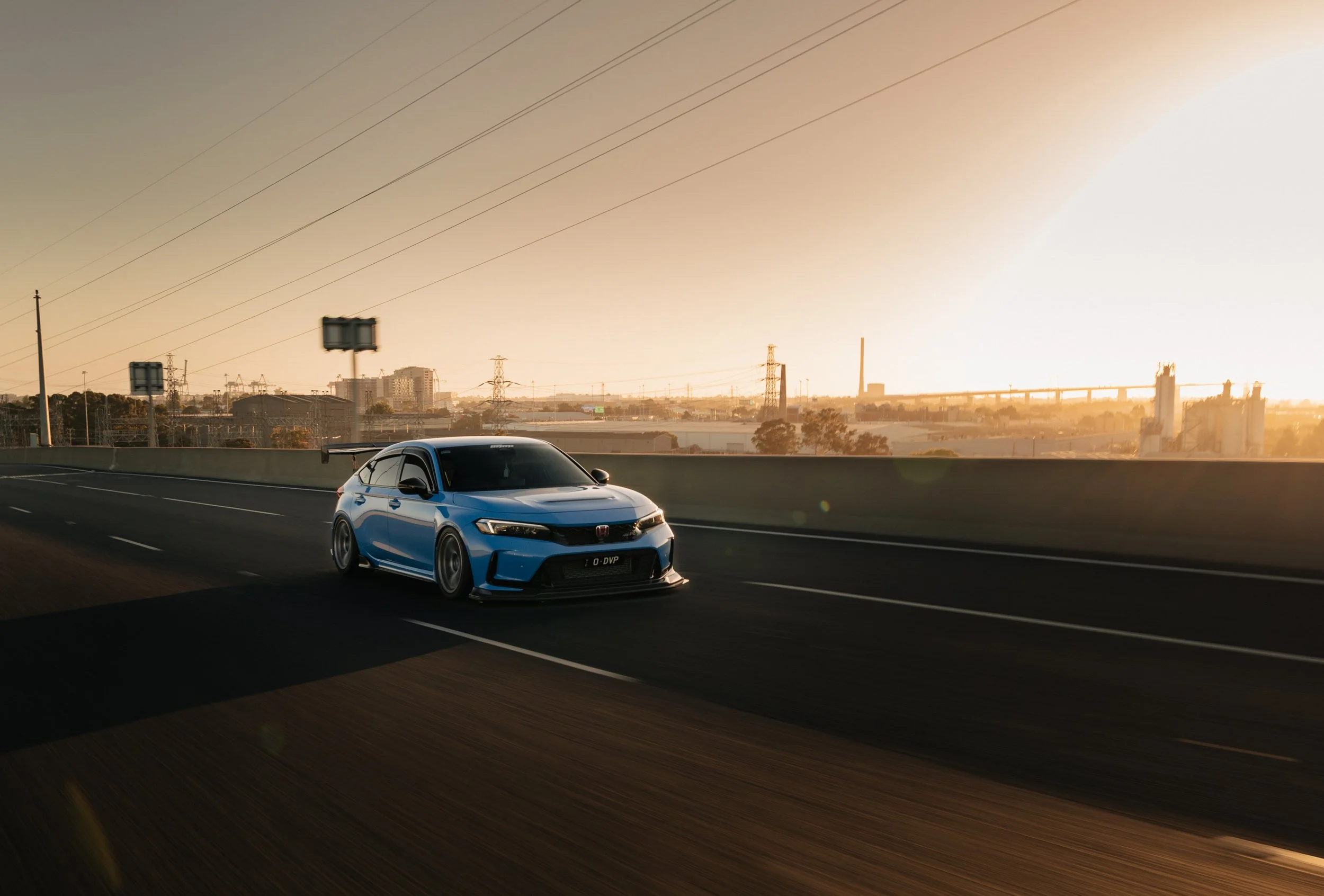 A blue Honda Civic driving on a highway during sunset with an industrial landscape and power lines in the background.