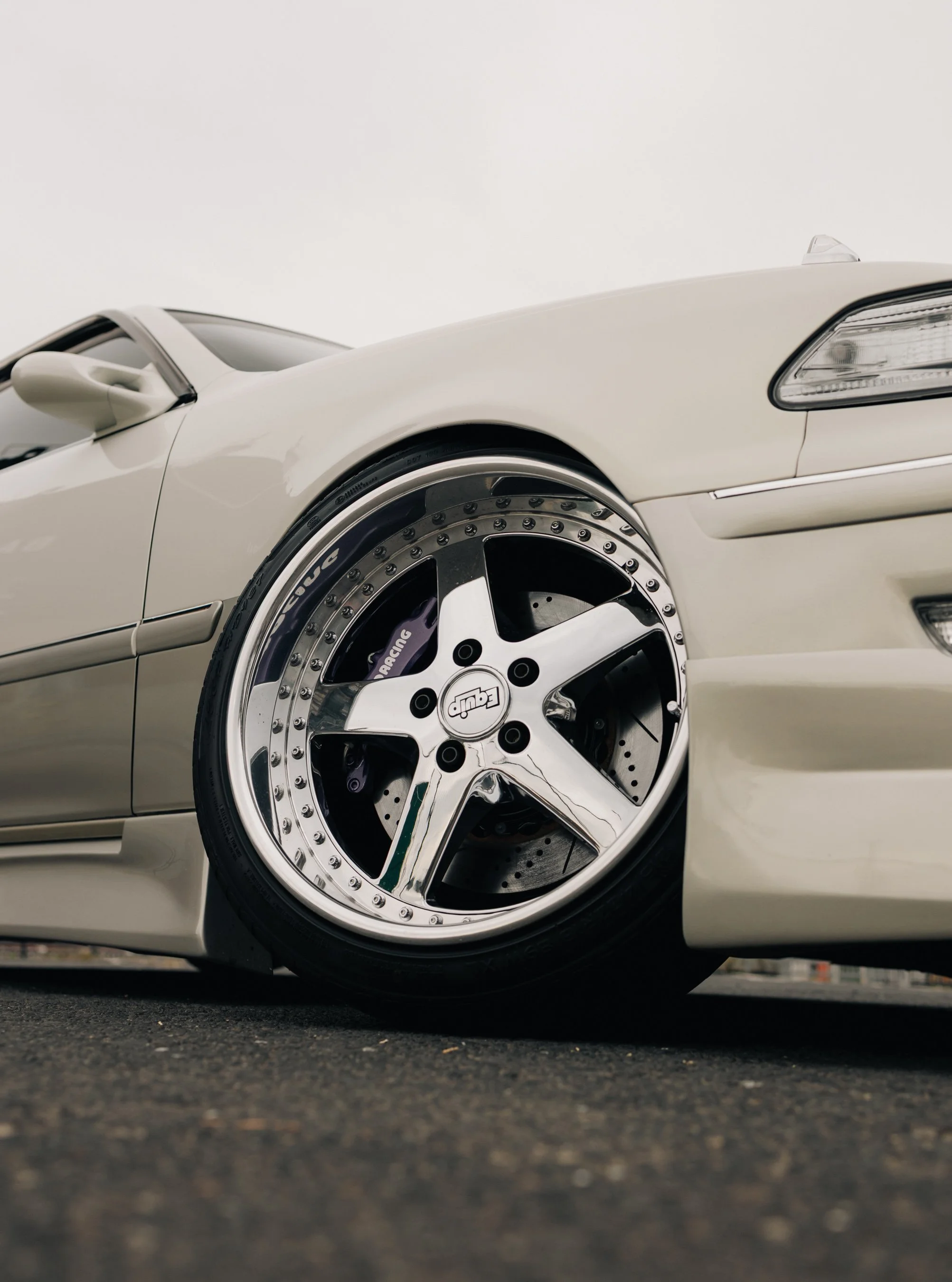 Close-up of a white sports car showing its front wheel with a chrome rim, brake disc, and caliper, on a paved surface with a plain sky background.