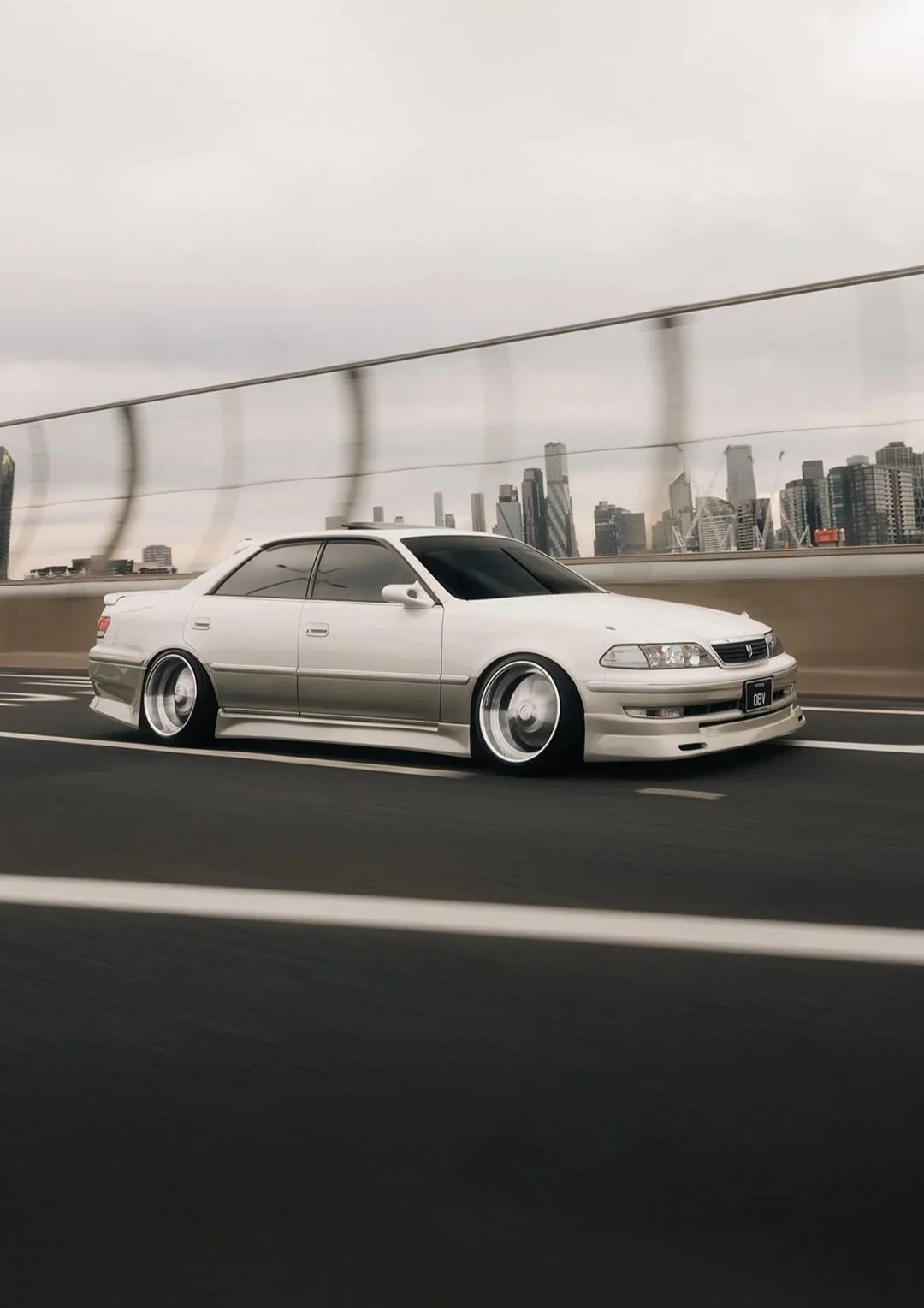 A white Acura sedan with modifications, including lowered suspension and aftermarket wheels, driving on a highway with a city skyline in the background.