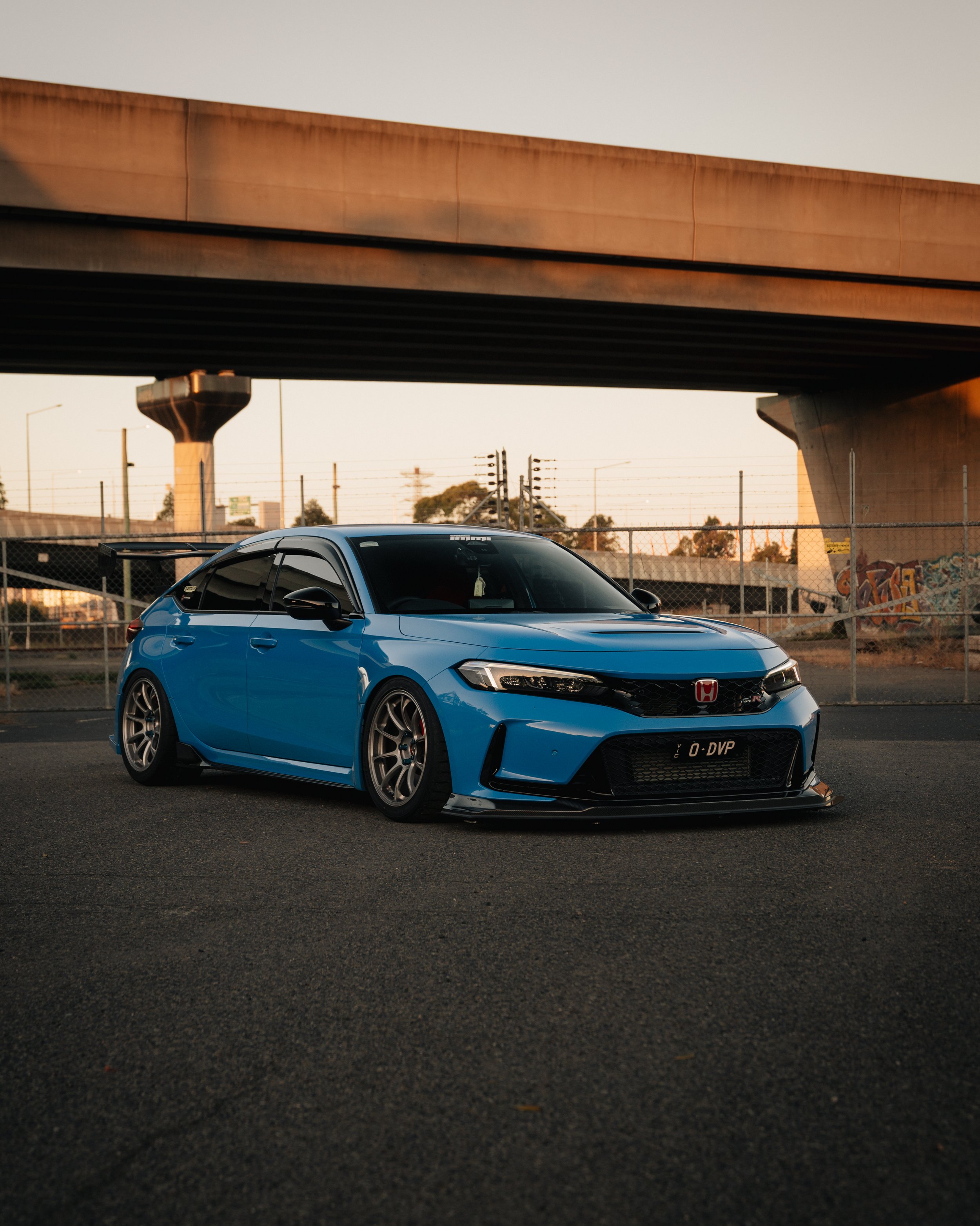 Blue Honda Civic parked under a bridge in an industrial area during sunset.