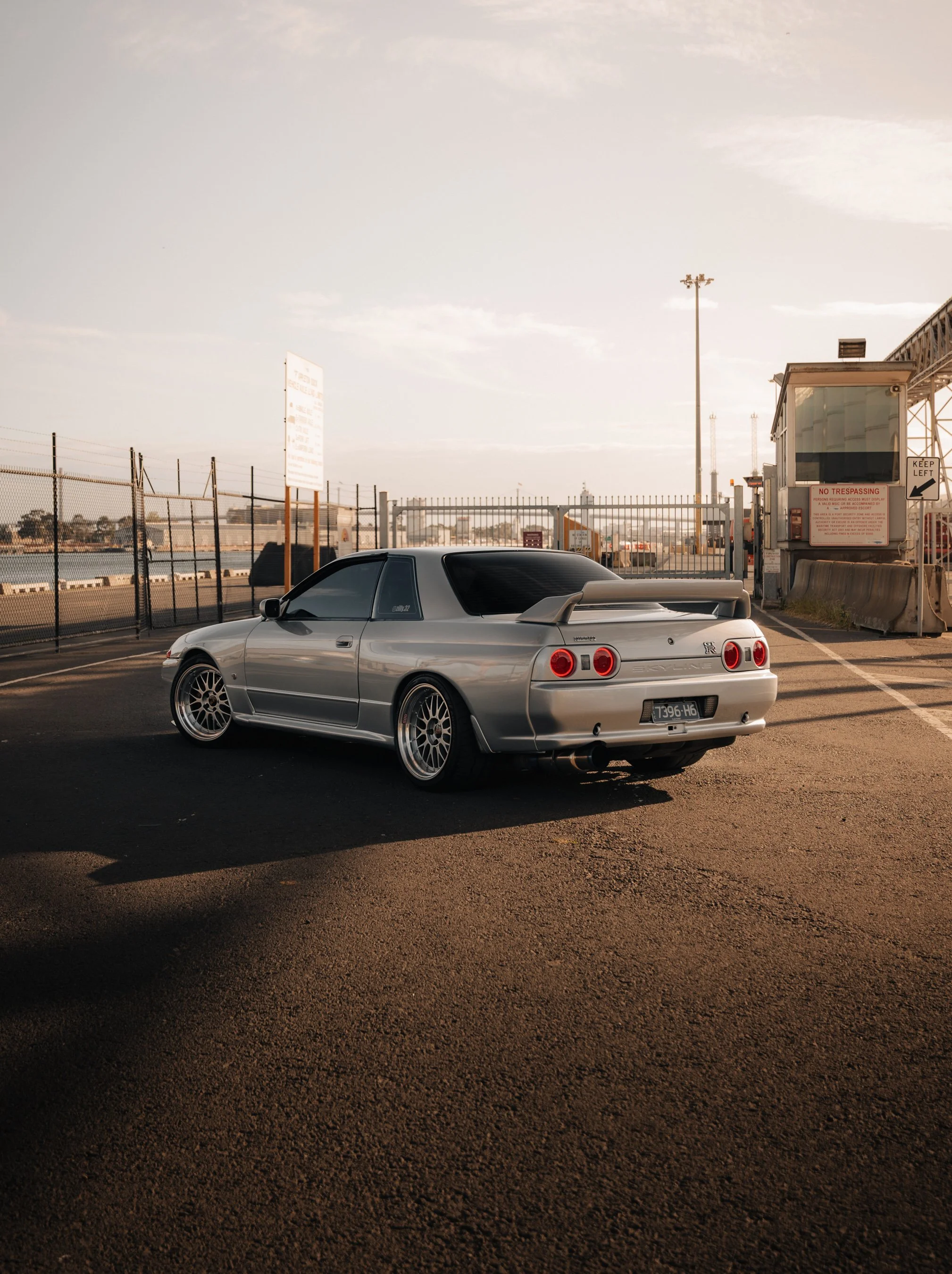 Silver Nissan Skyline GTR parked near a waterfront, with a chain-link fence and port security booth in the background during sunset.