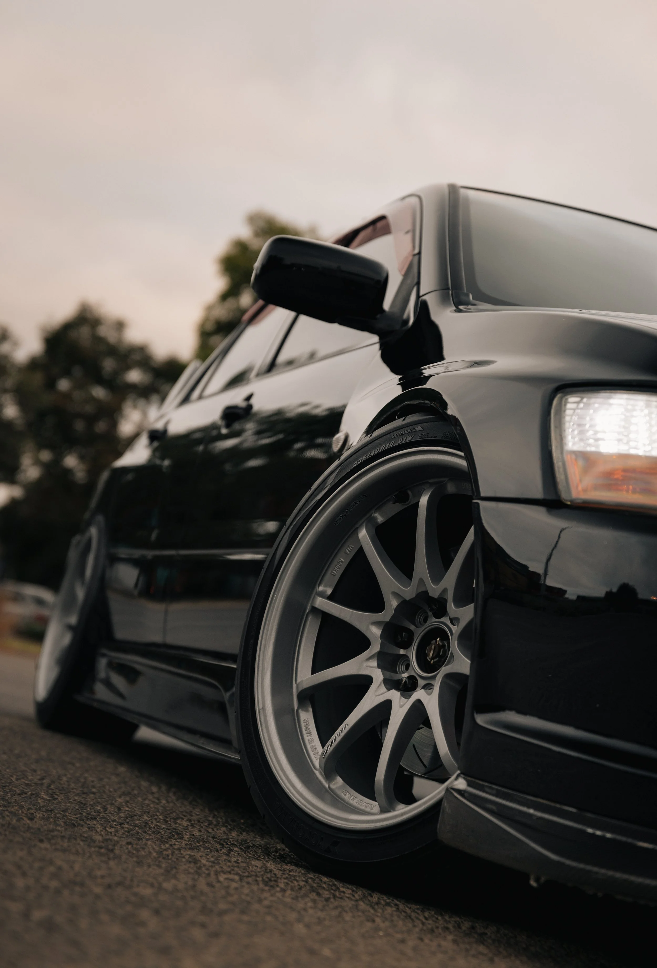 Close-up of a black car with silver custom wheels, taken from a low angle during dusk with a blurred background of trees and a cloudy sky.