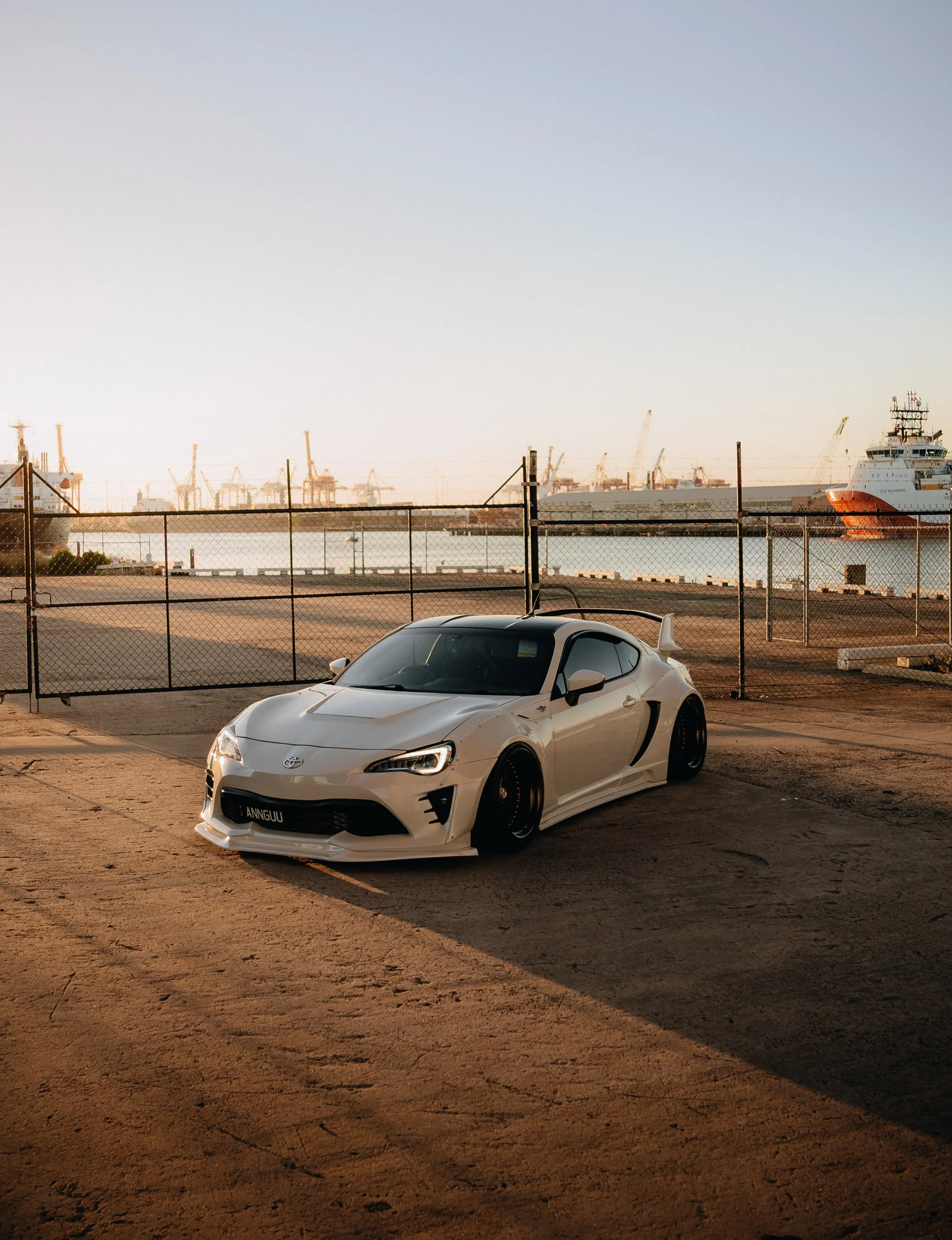 A white sports car with a custom license plate reading 'ANNGUU' parked near a chain-link fence at the harbor during sunset, with large ships and cranes in the background.