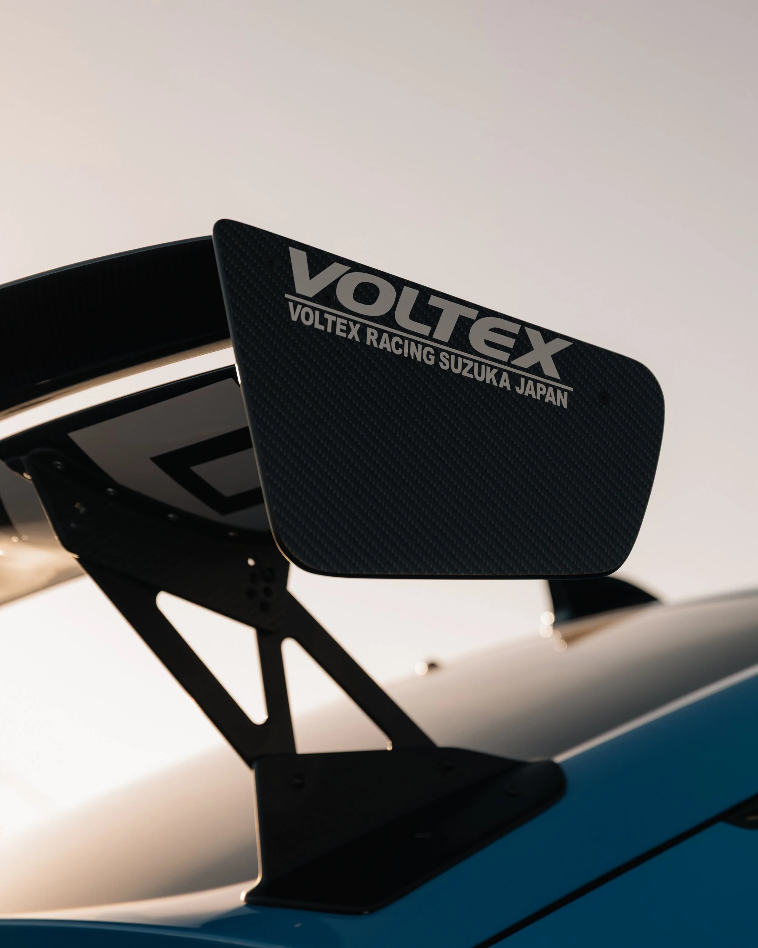 Close-up of a black racing car rear wing with the words "VOLTEQ VOLTEX RACING SUZUKA JAPAN" written on it, against a bright sky background.