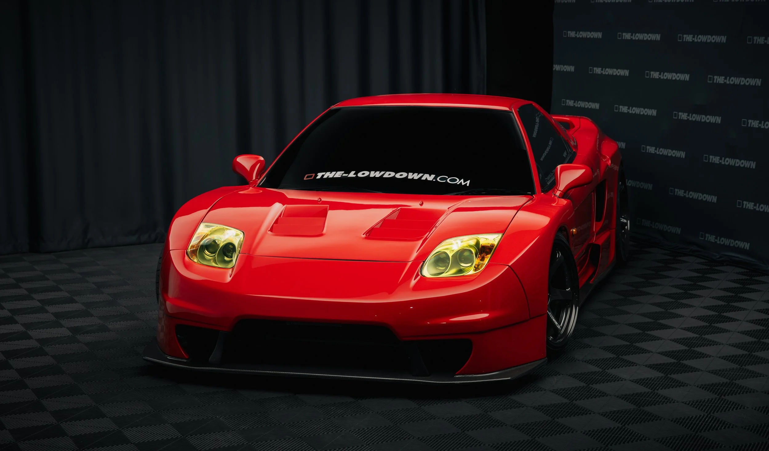 A red sports car with yellow-tinted headlights displayed indoors, with a backdrop featuring 'THE-LOWDOWN' branding.