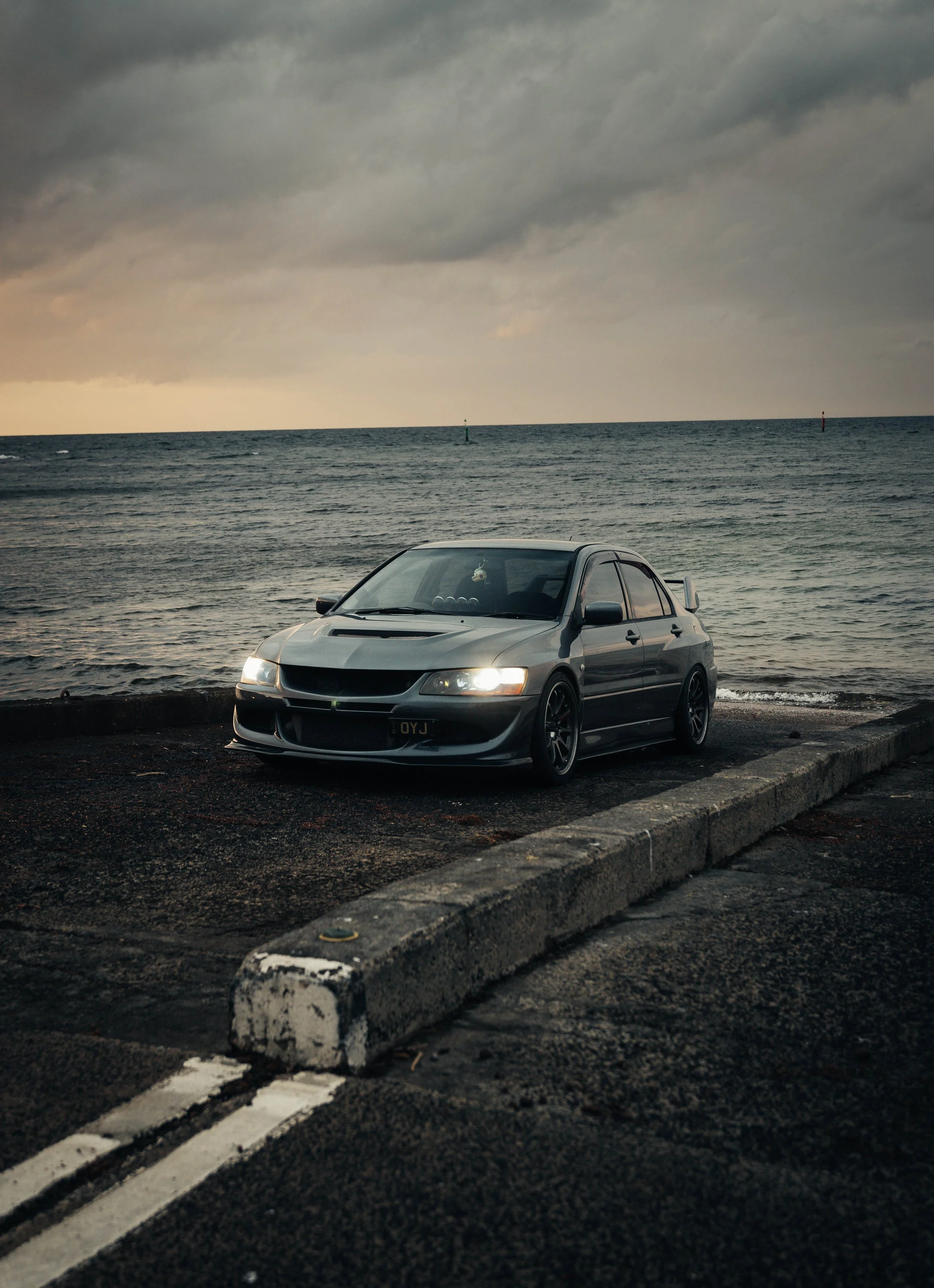 A black modified car parked on a concrete lot by the ocean during cloudy weather, with the sea and a cloudy sky in the background. EVO 8, evo 9 Mitshubishi evolution, jdm car, jdm photo, work wheels, work racing, race car, speed, jdm. 