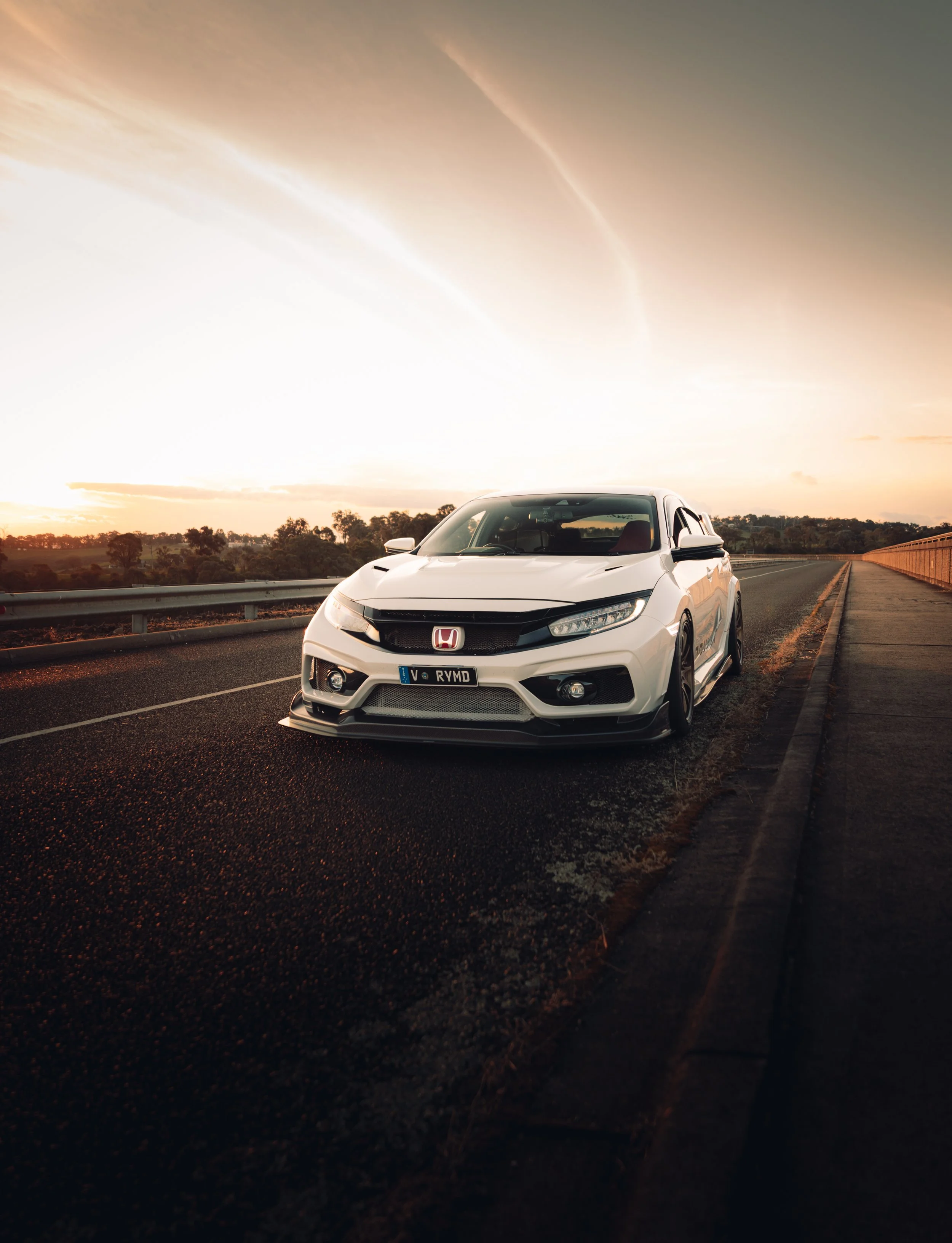 A white Honda Civic car parked on the side of a highway during sunset with a scenic background of trees and sky.