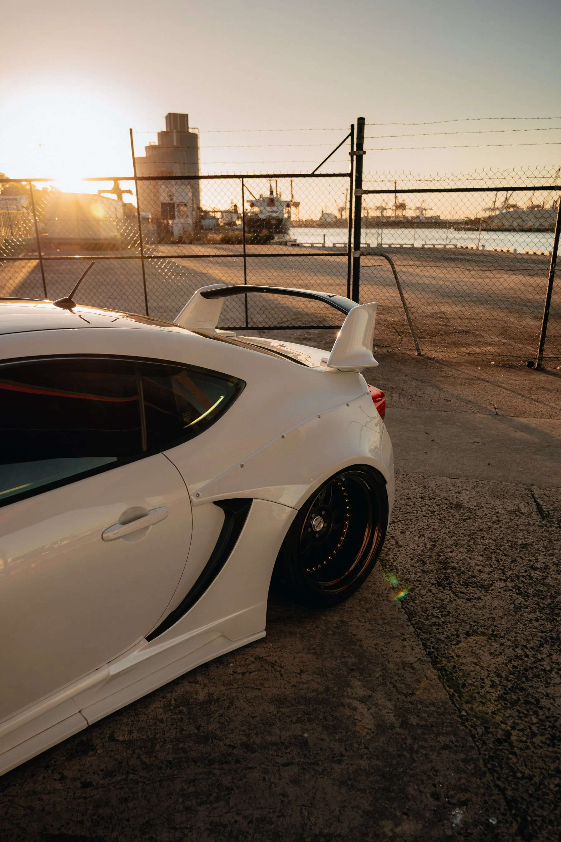 White sports car with a large rear spoiler parked near the water at sunset, with ships and industrial buildings in the background. White Toyota 86 photo, sunset time, golden hour, widebody 86, work wheels, low car, jdm.