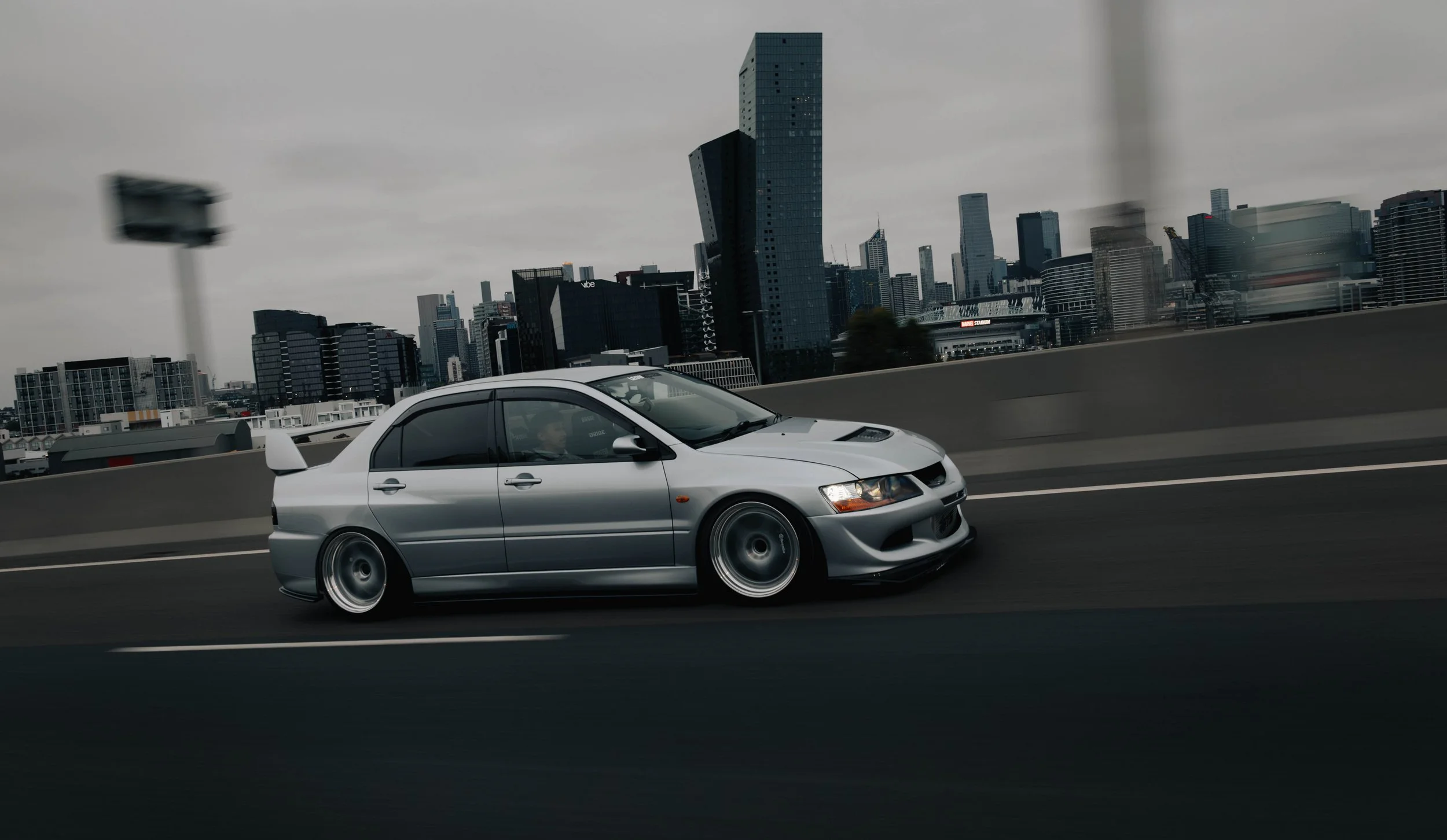 A silver sports car is driving on a highway with a city skyline in the background, under a cloudy sky.