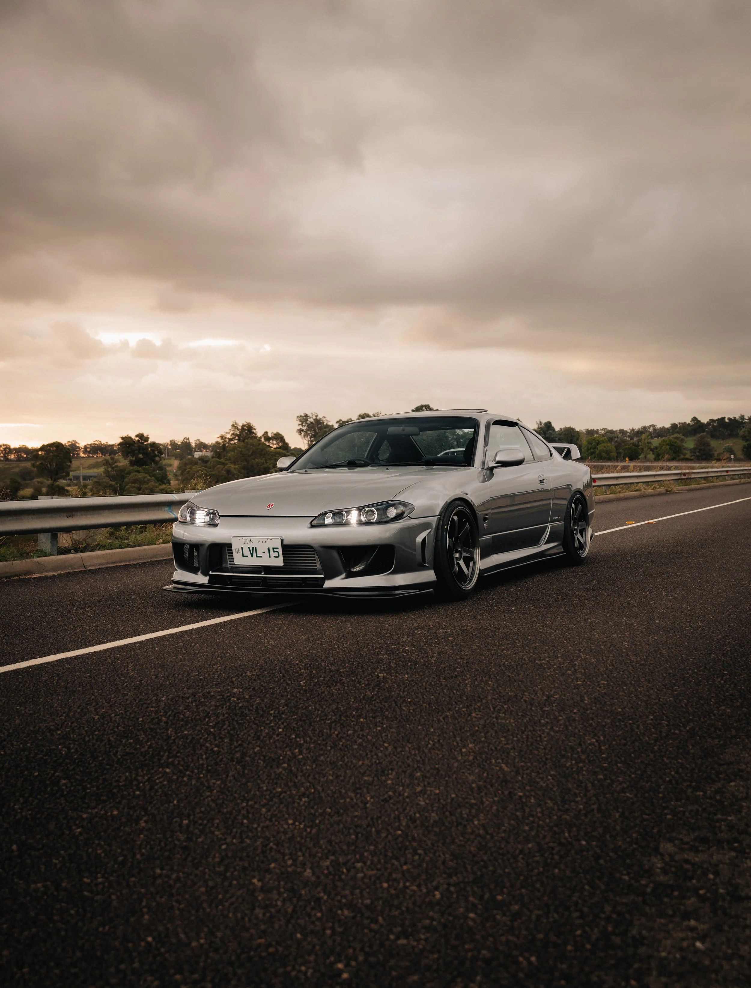 A silver sports car parked on the side of a road under a cloudy sky, with trees and hills in the background.