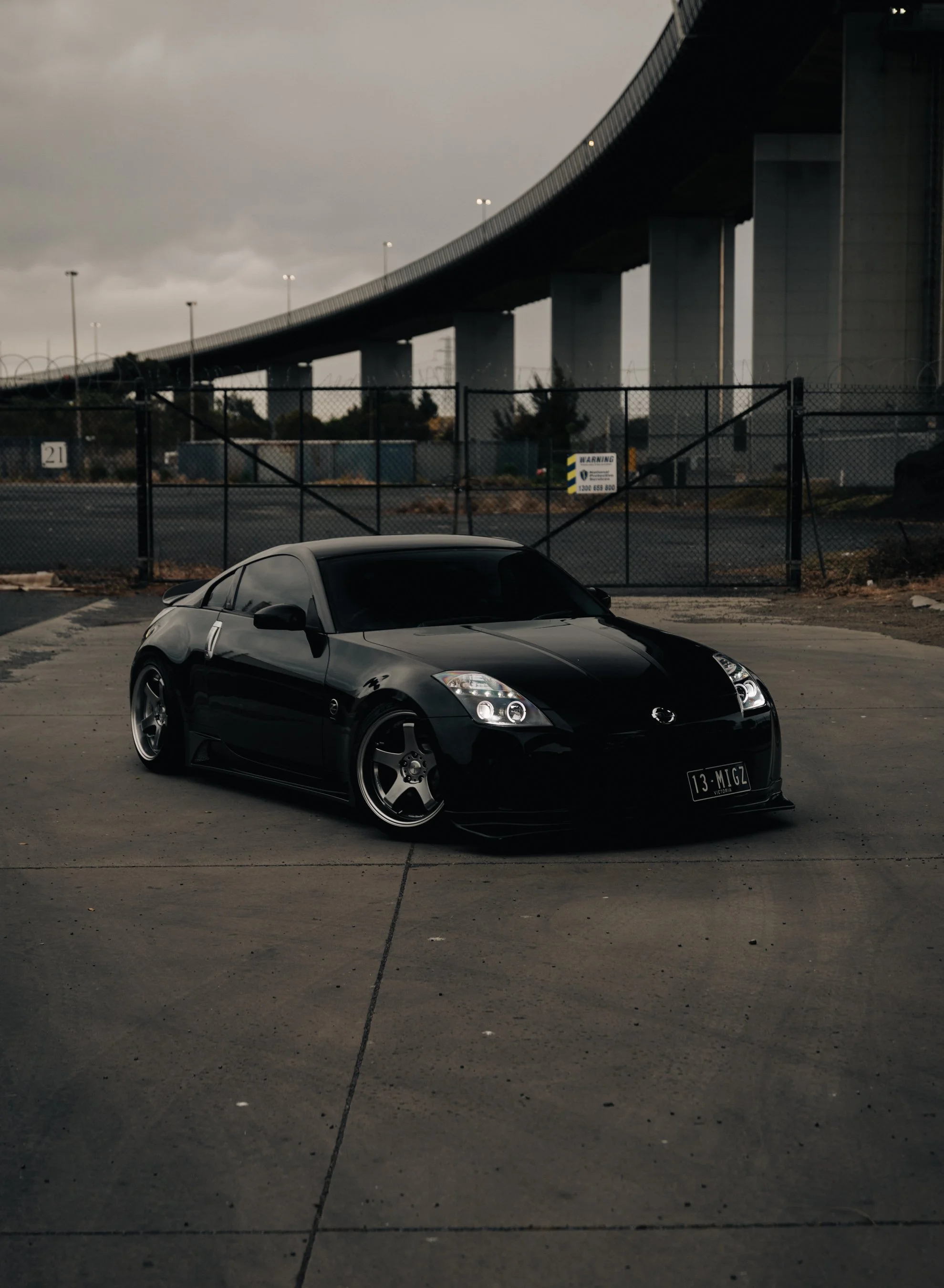 A black sports car parked on a concrete surface beneath a large highway bridge, with a chain-link fence and overcast sky in the background.