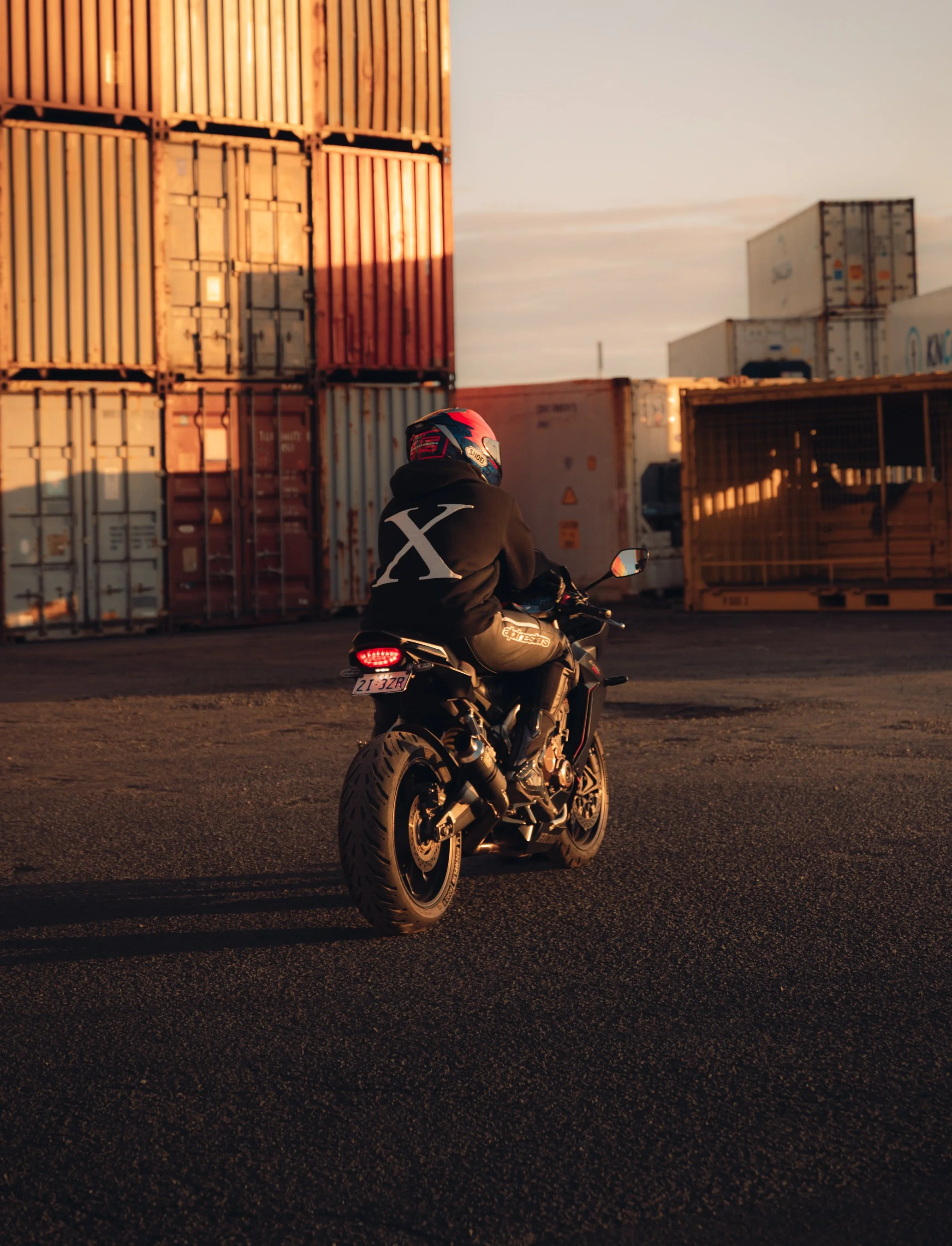 Motorcyclist riding a black motorcycle near shipping containers during sunset.