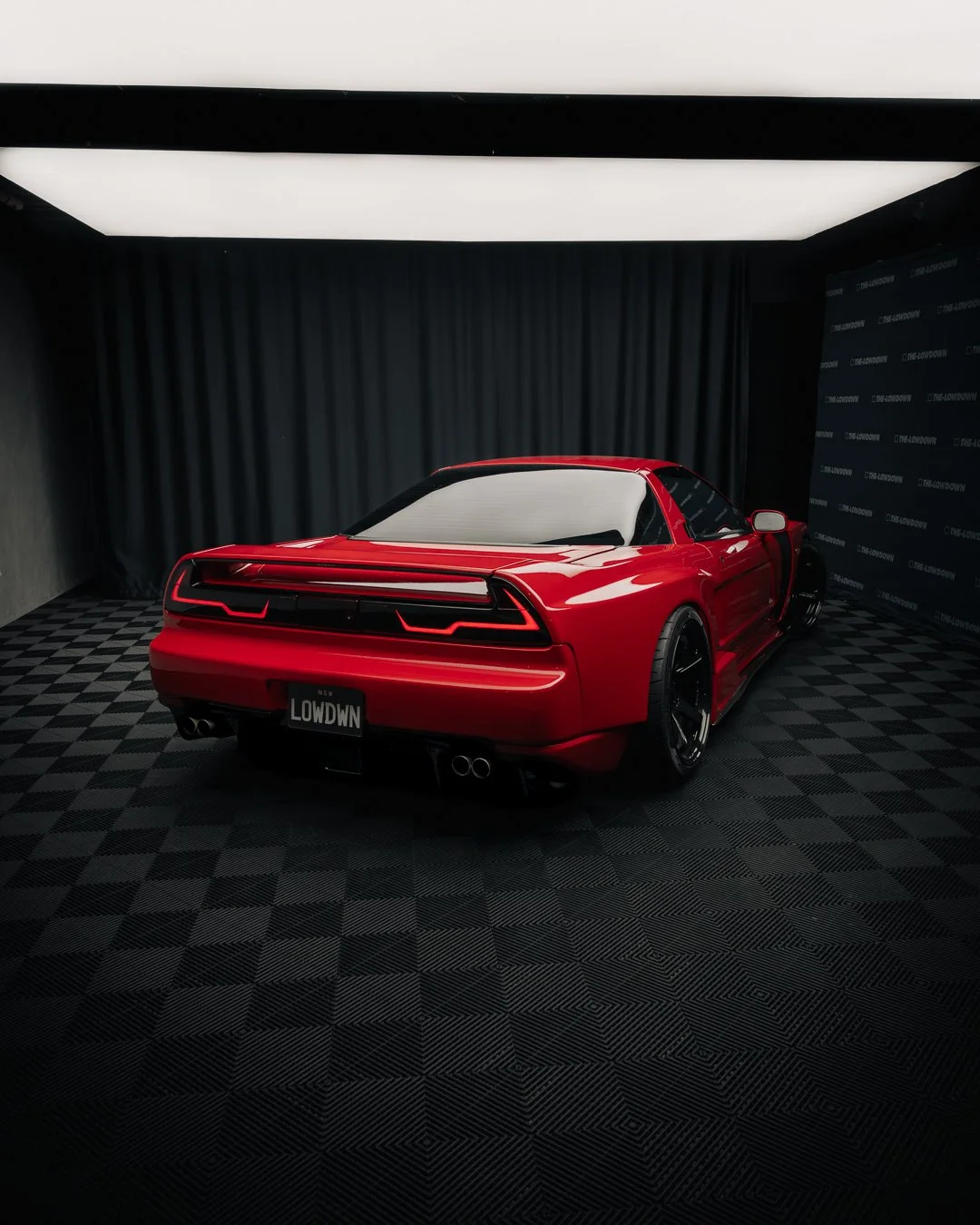 Red sports car with black accents displayed in a showroom or studio with black curtains and a checkered floor.