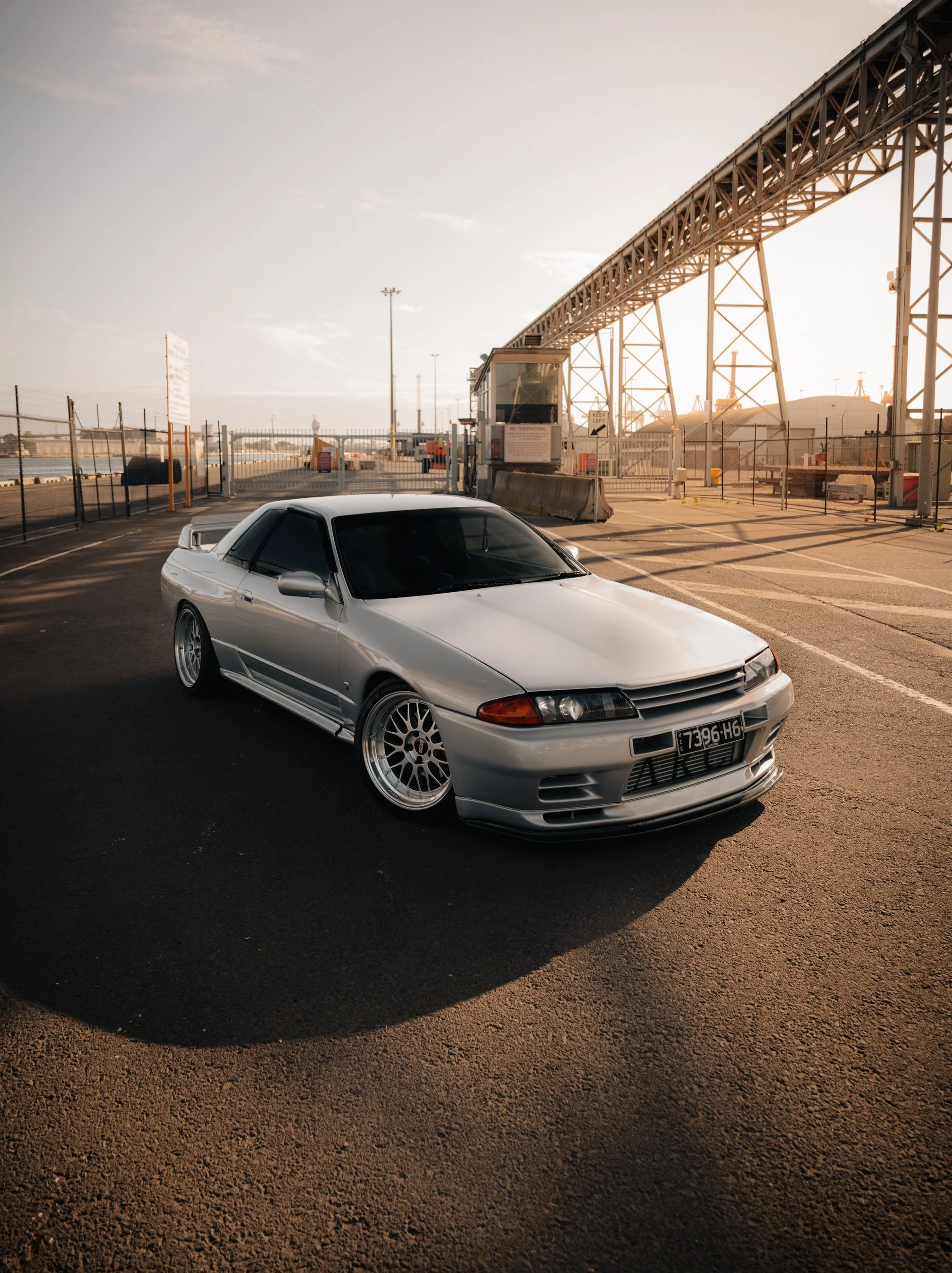 Silver sports car parked in an industrial area at sunset, with a chain-link fence, a guard booth, and a large metal structure overhead. Nissan R32 GTR Skyline, GTR photo, dock photo, car photos, jdm at the docks, melbourne cars, melbourne docks, BBS 