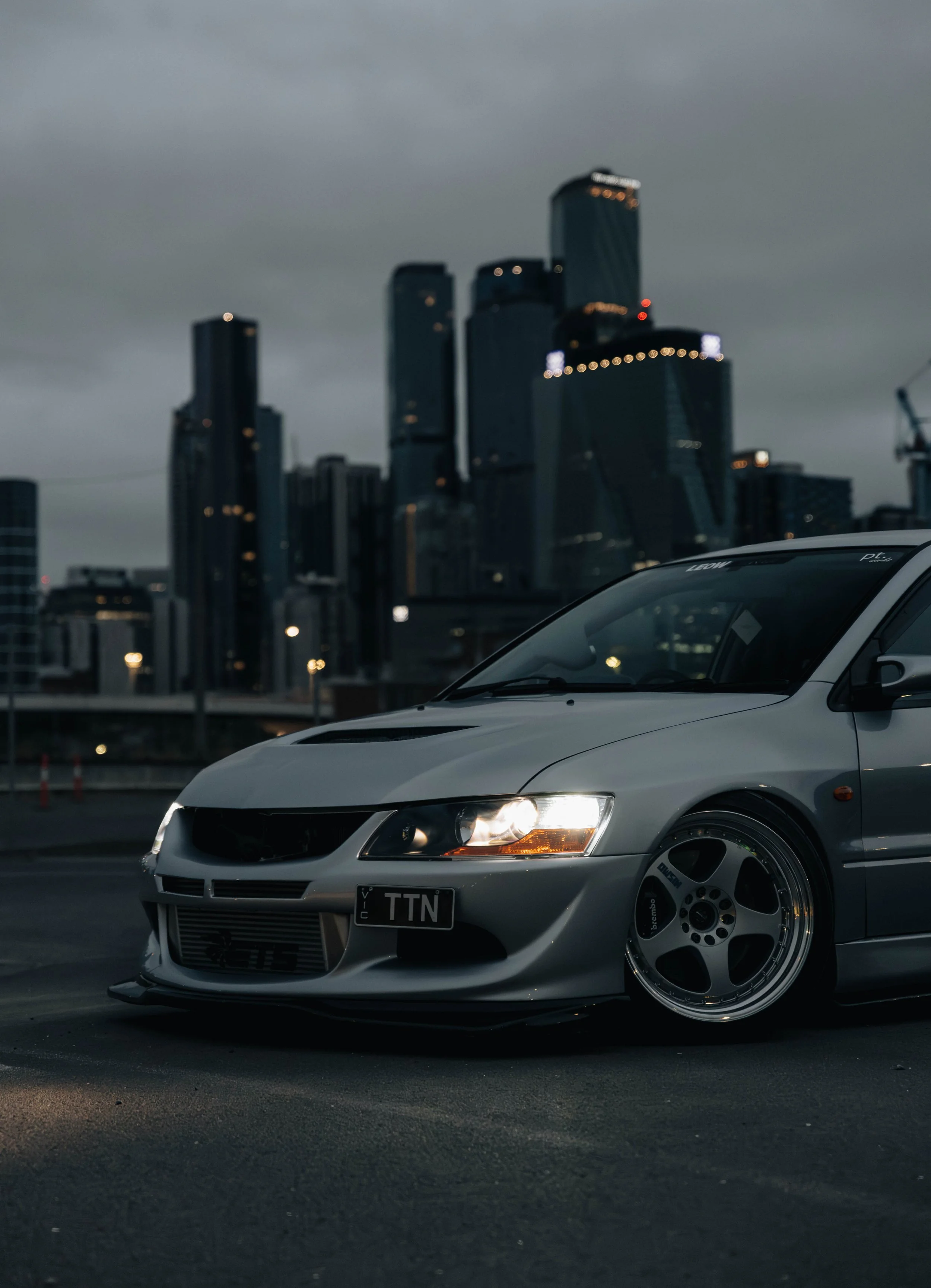 A silver sports car with modified wheels and lowered suspension parked on a dark asphalt surface, with a city skyline of tall buildings and cloudy sky in the background.