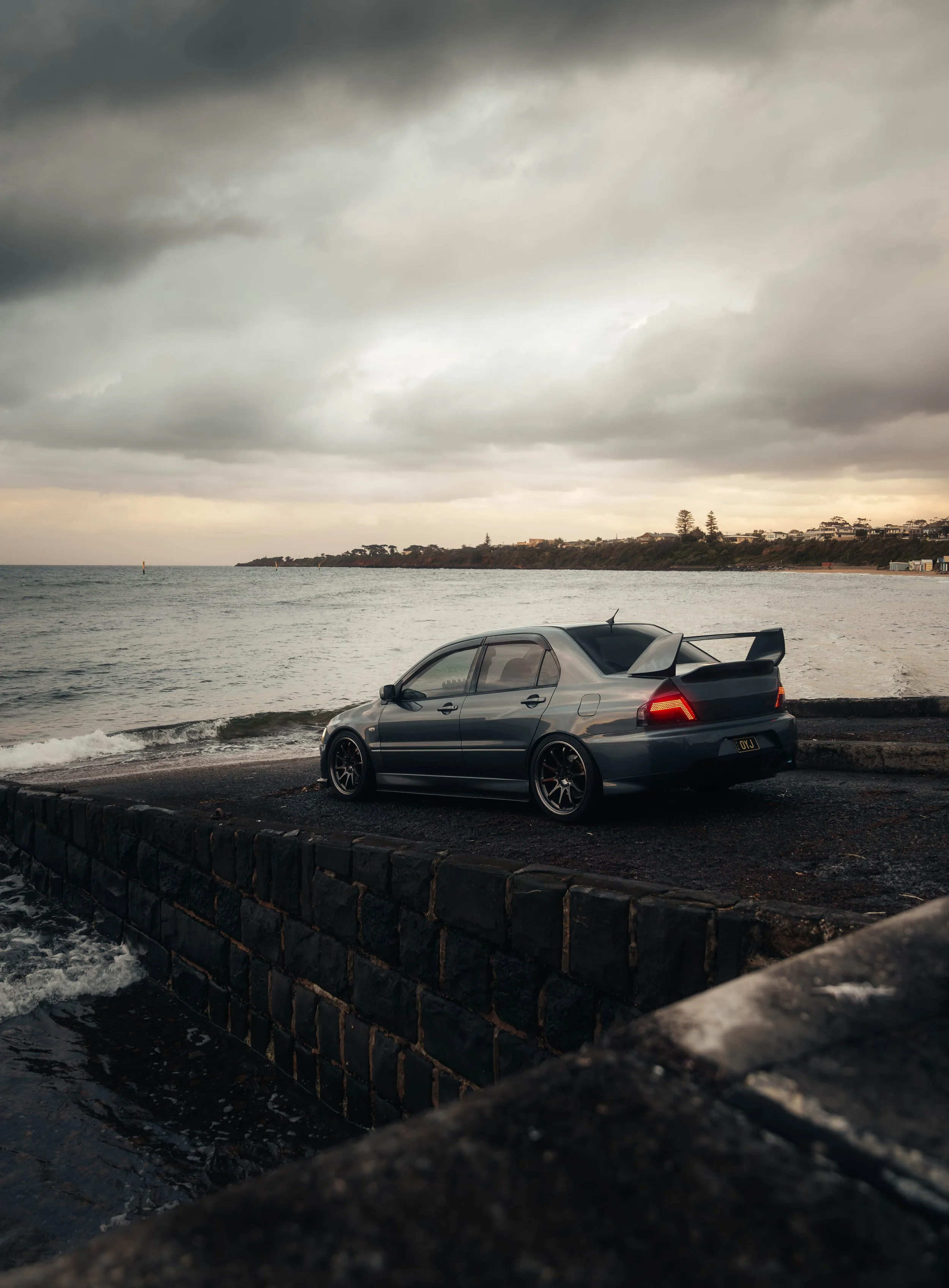 A dark gray sports car with a large rear spoiler parked on a small ferry dock beside the ocean, under a cloudy sky, with a rocky wall in the foreground and a distant shoreline in the background. EVO 8, evo 9 Mitshubishi evolution, jdm car, jdm photo,