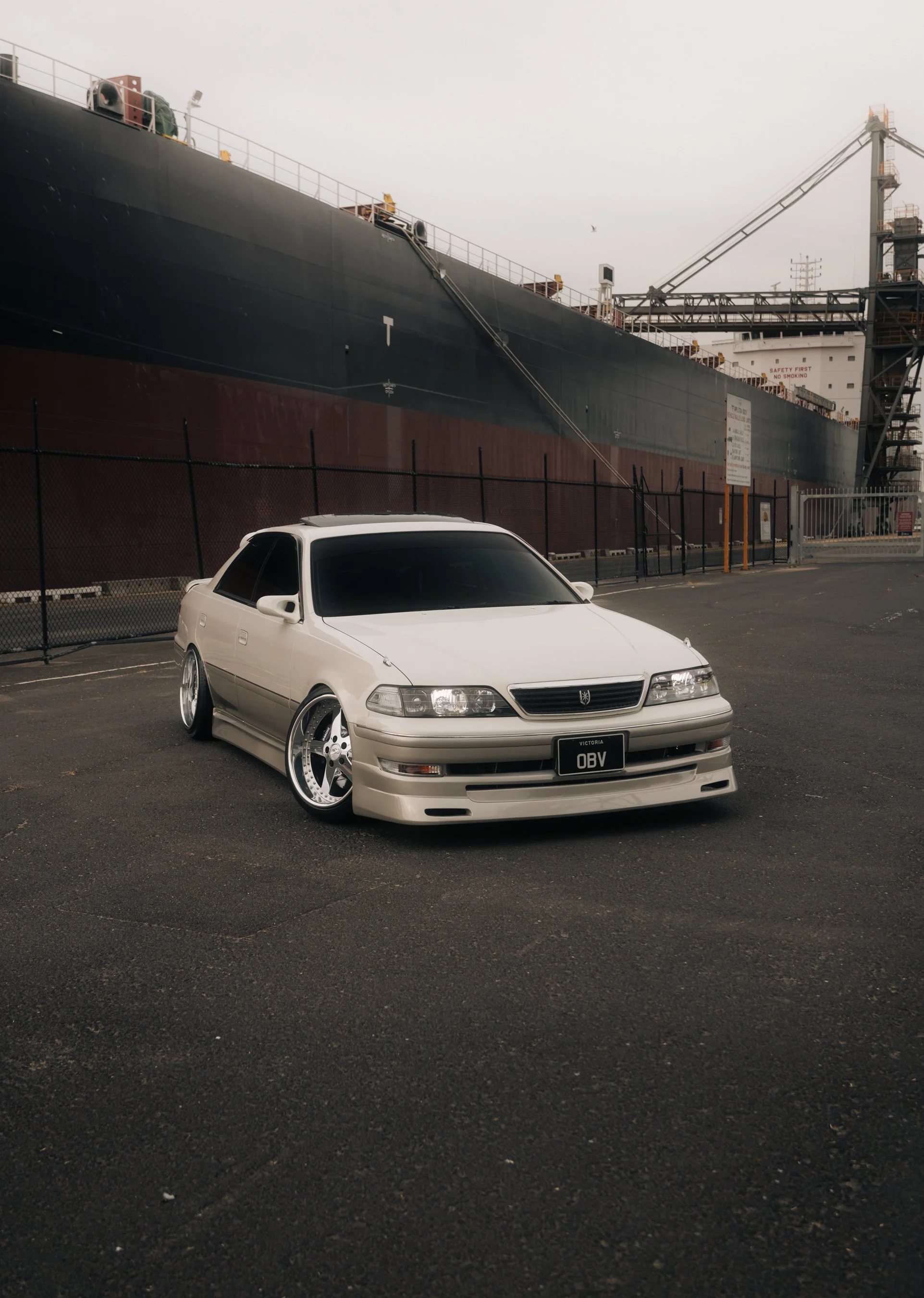 A white modified Honda Accord with lowered suspension and custom wheels parked in an empty parking lot near a large cargo ship docked at a port.