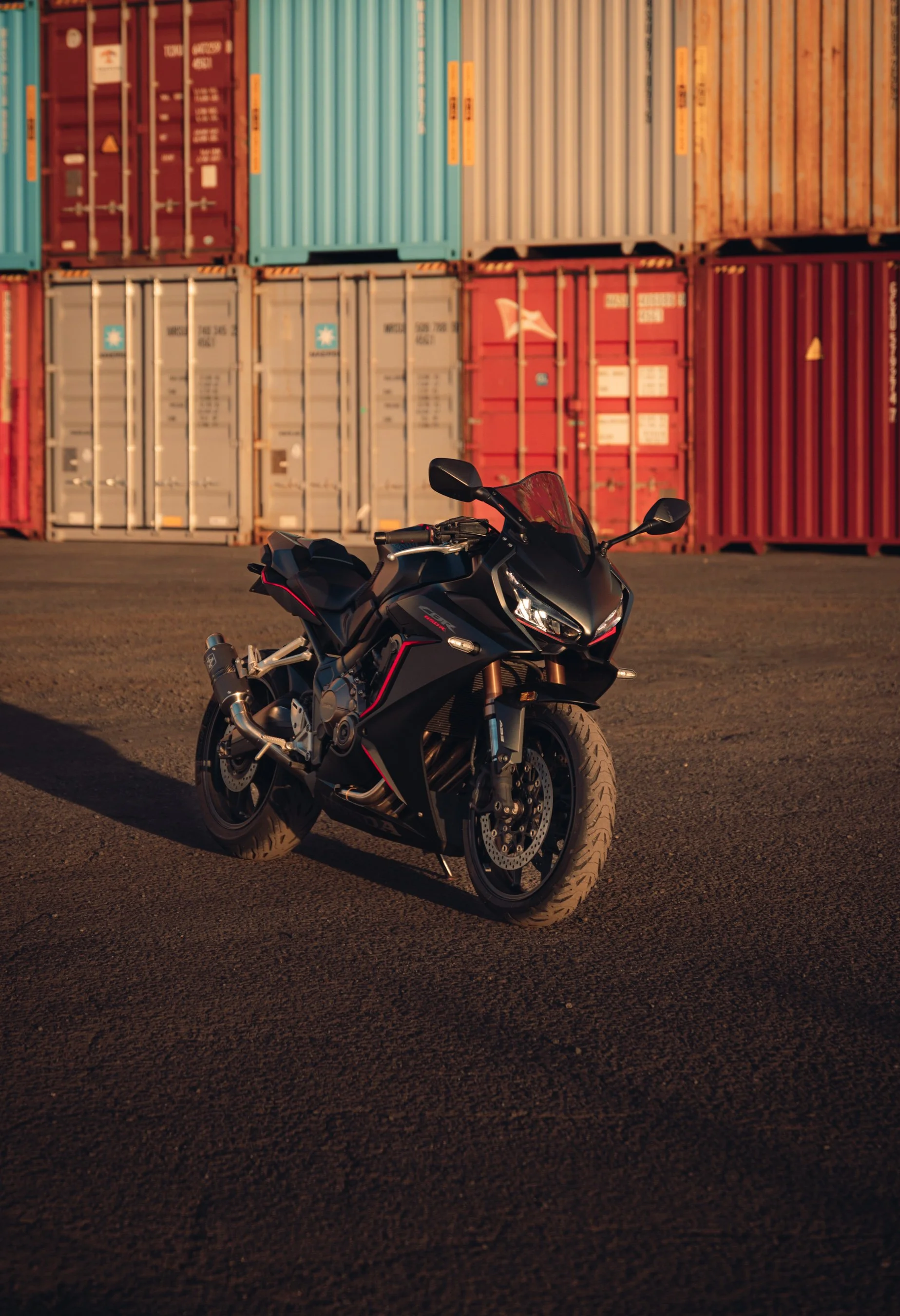 A black sport motorcycle parked on an asphalt surface with colorful shipping containers in the background at sunset.