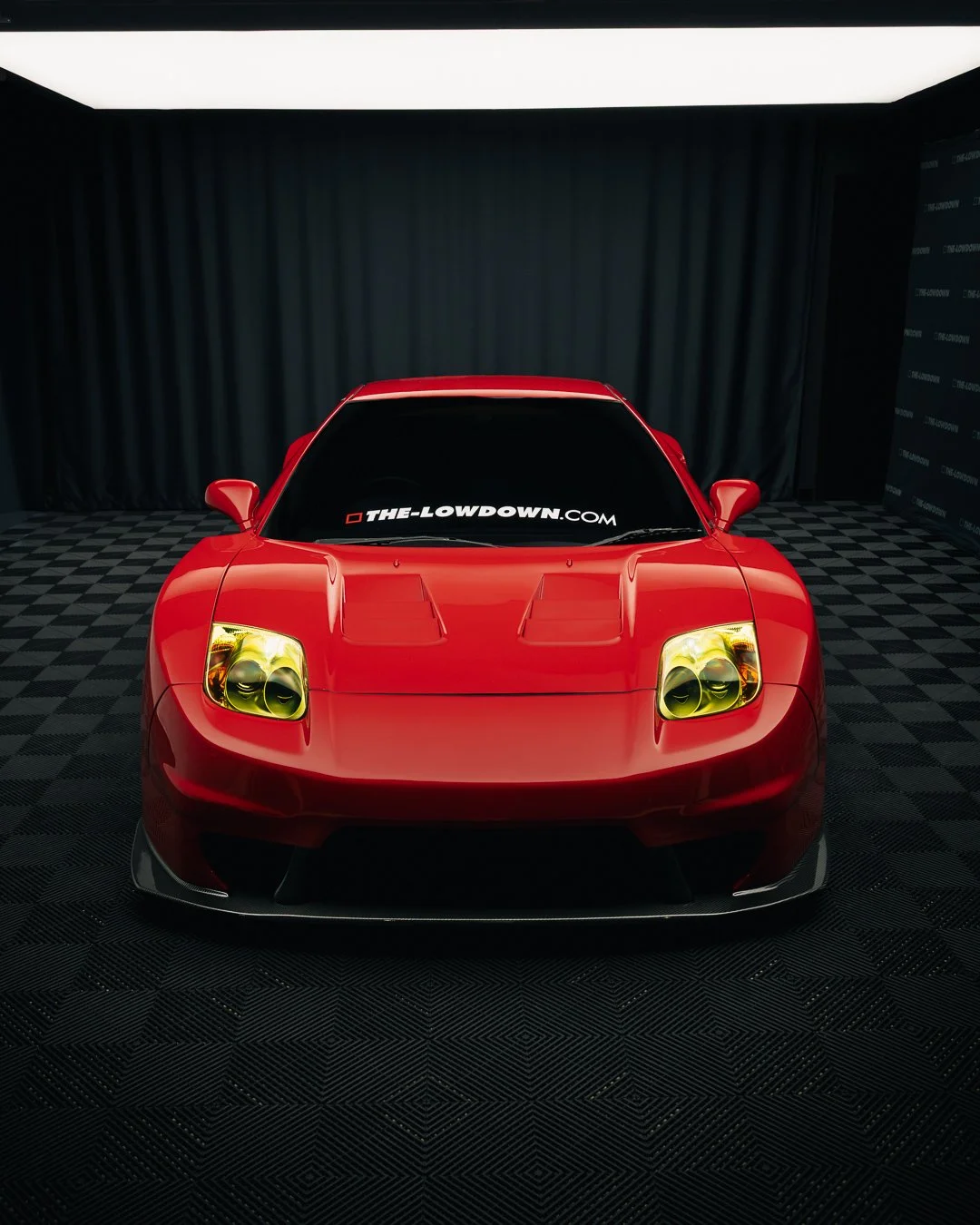 Red sports car with yellow-tinted headlights, black tinted windows, and a flat front hood, parked indoors with checkered flooring and black curtains.