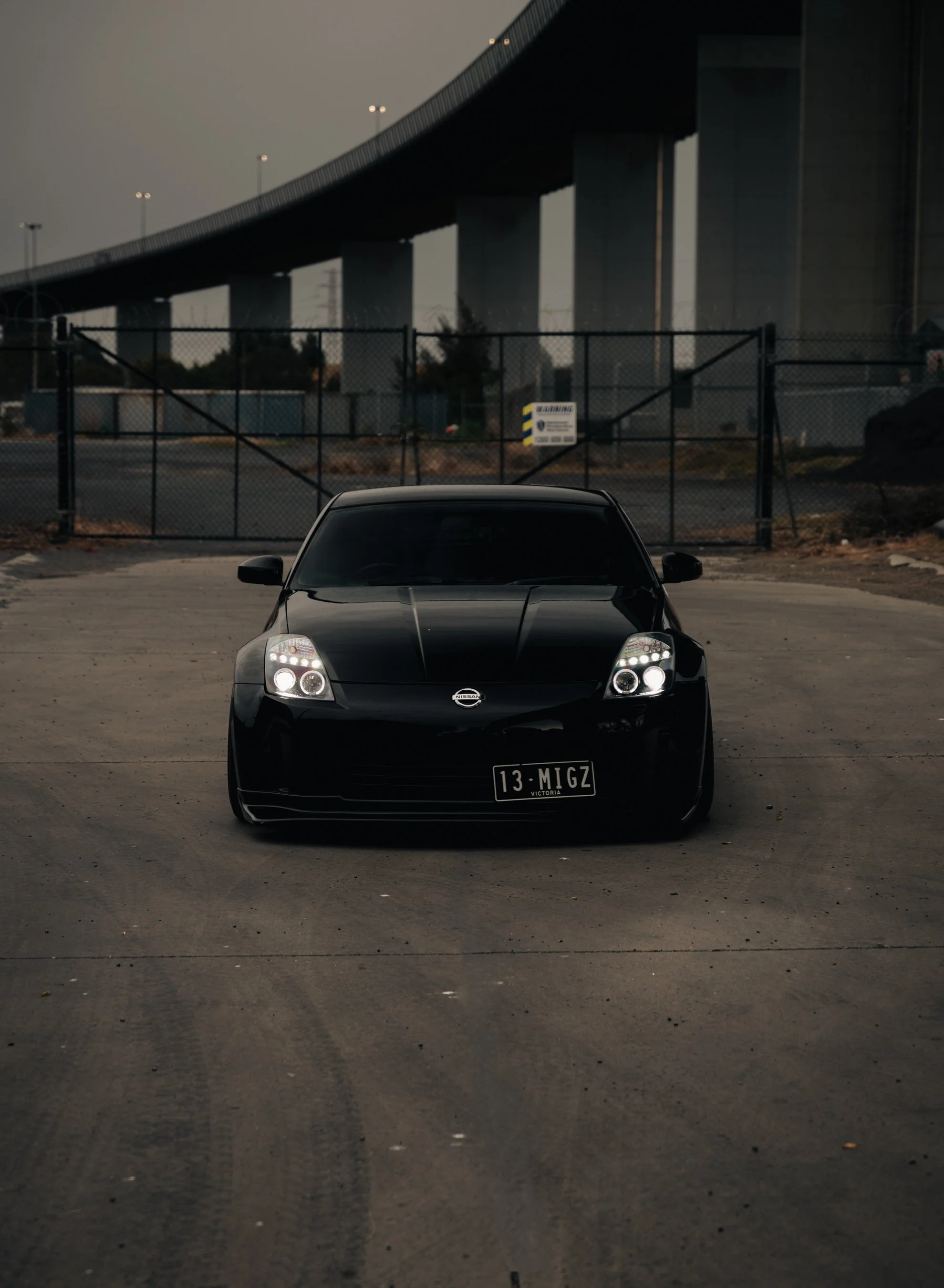 Black Nissan 350Z sports car parked in an empty lot, with a large bridge overhead and a fence in the background. Nissan 350Z, jdm, nissan photo, jdm car, work wheels, work photo, underground spot, lowered car. 