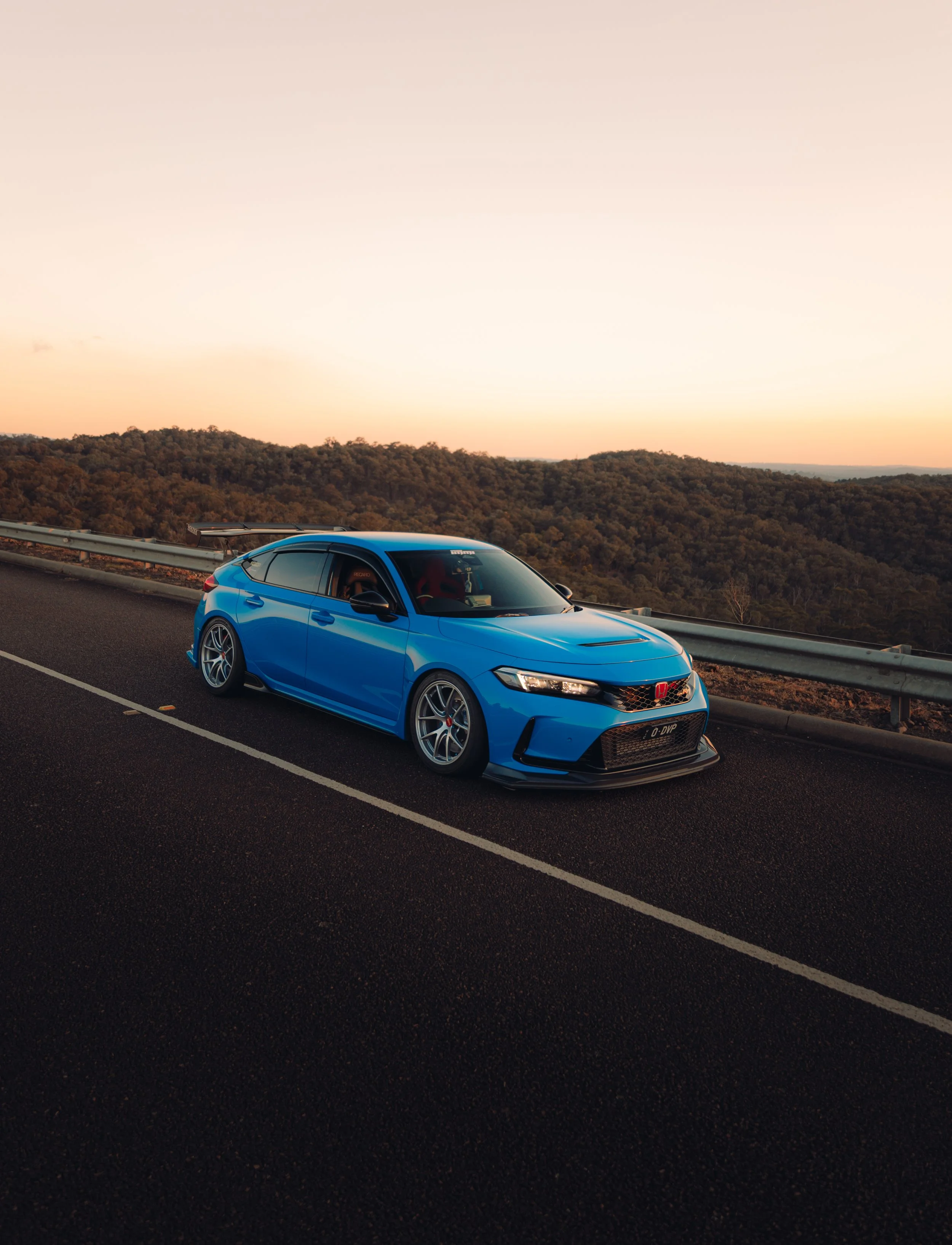 A blue Honda Civic type R parked on the side of a mountain road at sunset, overlooking a forested landscape.