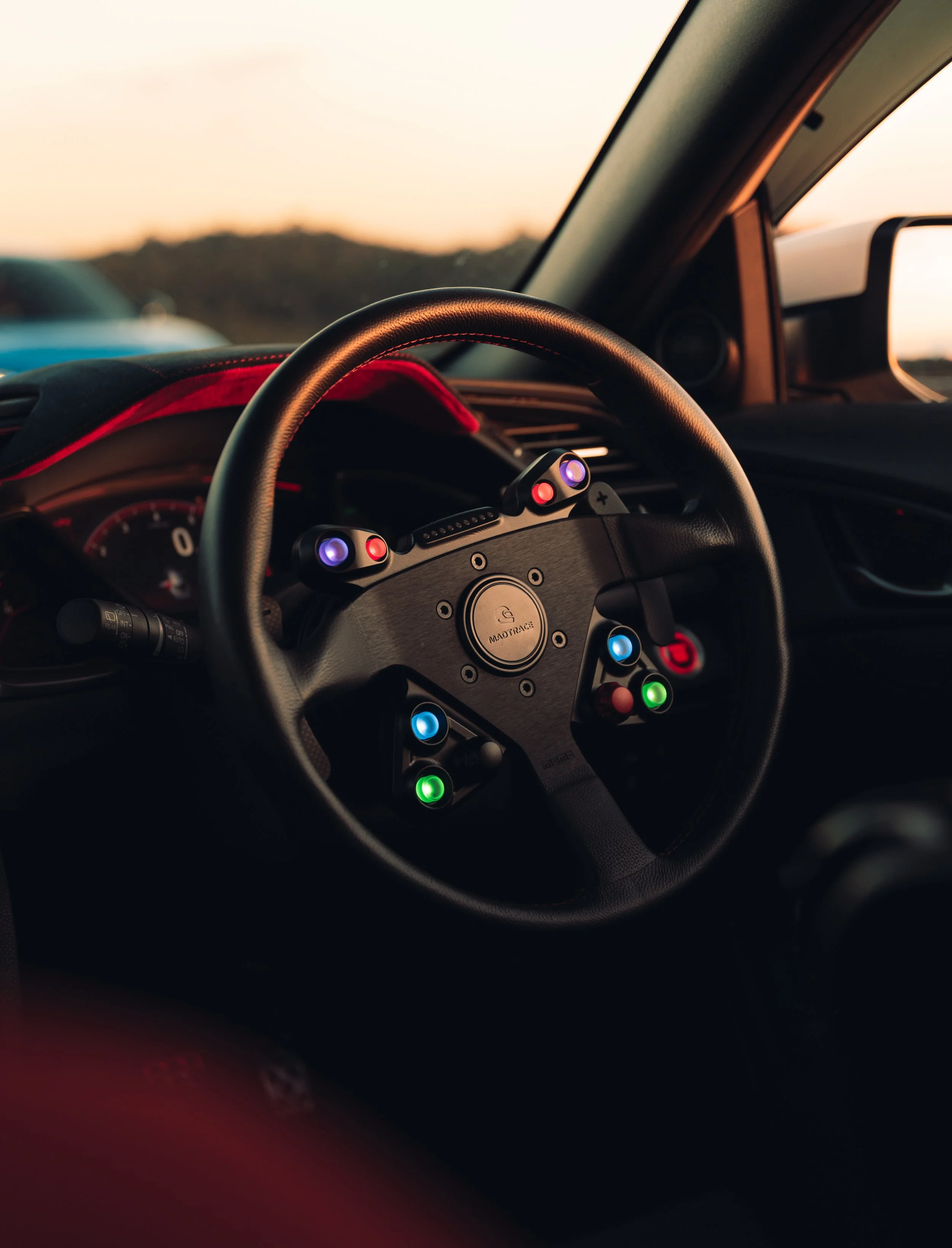 Close-up of a racing car steering wheel with colorful buttons, mounted on a dashboard with a speedometer and a sunset view through the windshield.