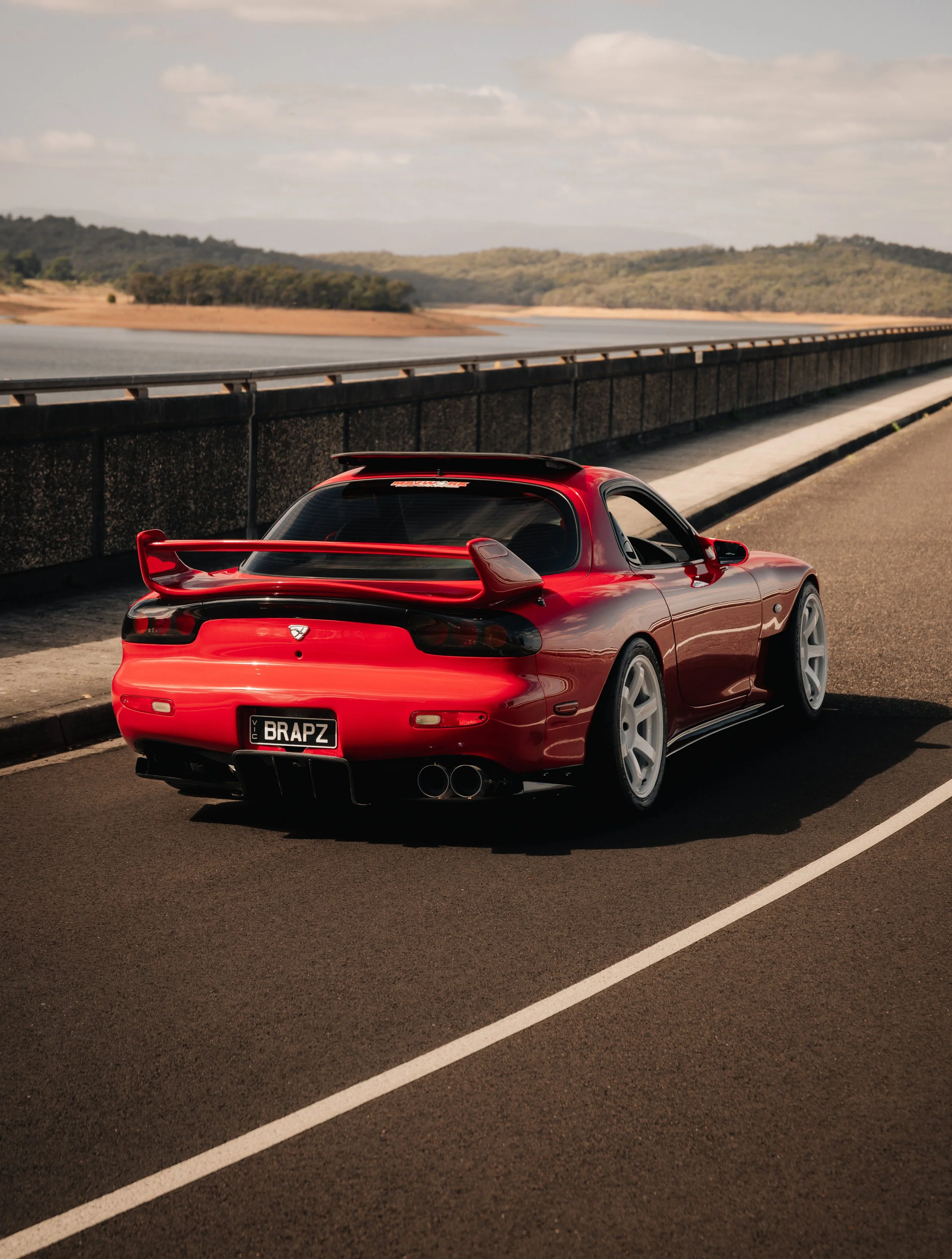 A red sports car with a large rear wing and custom license plate reading 'BRAPZ' parked on the side of a road next to a body of water and hilly landscape in the background.