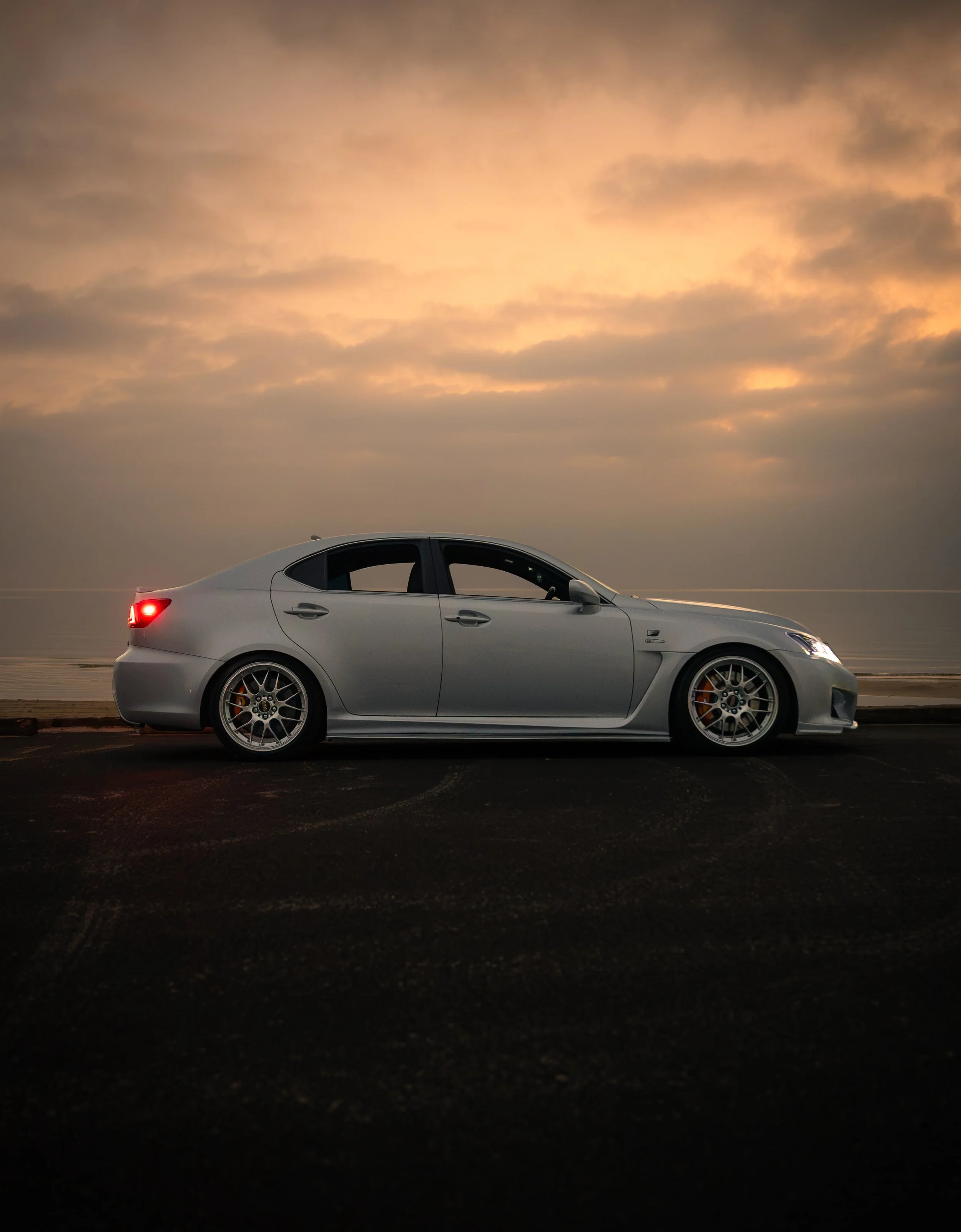 A silver car parked on a flat surface near the sea during sunset, with cloudy sky and soft light