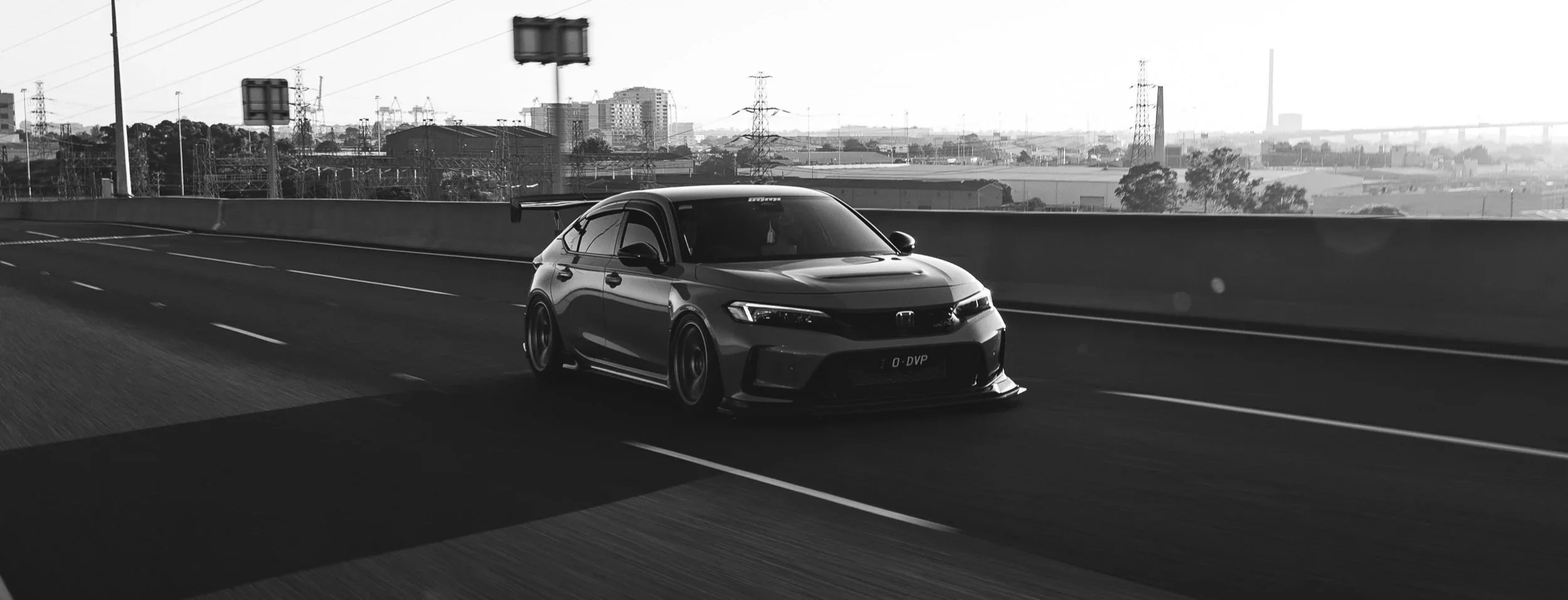A black and white photo of a sporty fl5 type r civic driving on a highway with city buildings, power lines, and a bridge in the background.