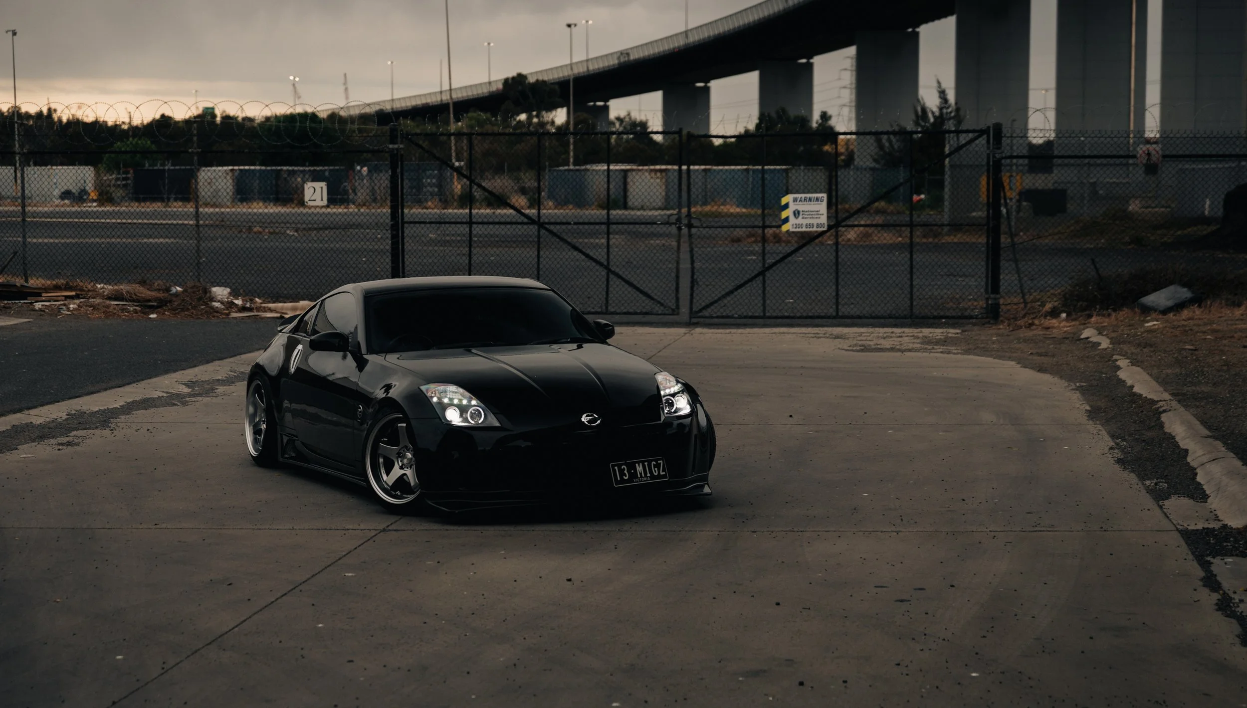 A black sports car parked on an empty concrete lot with a fence and industrial background, during dusk or dawn. Nissan 350Z, jdm, nissan photo, jdm car, work wheels, work photo, underground spot, lowered car. 