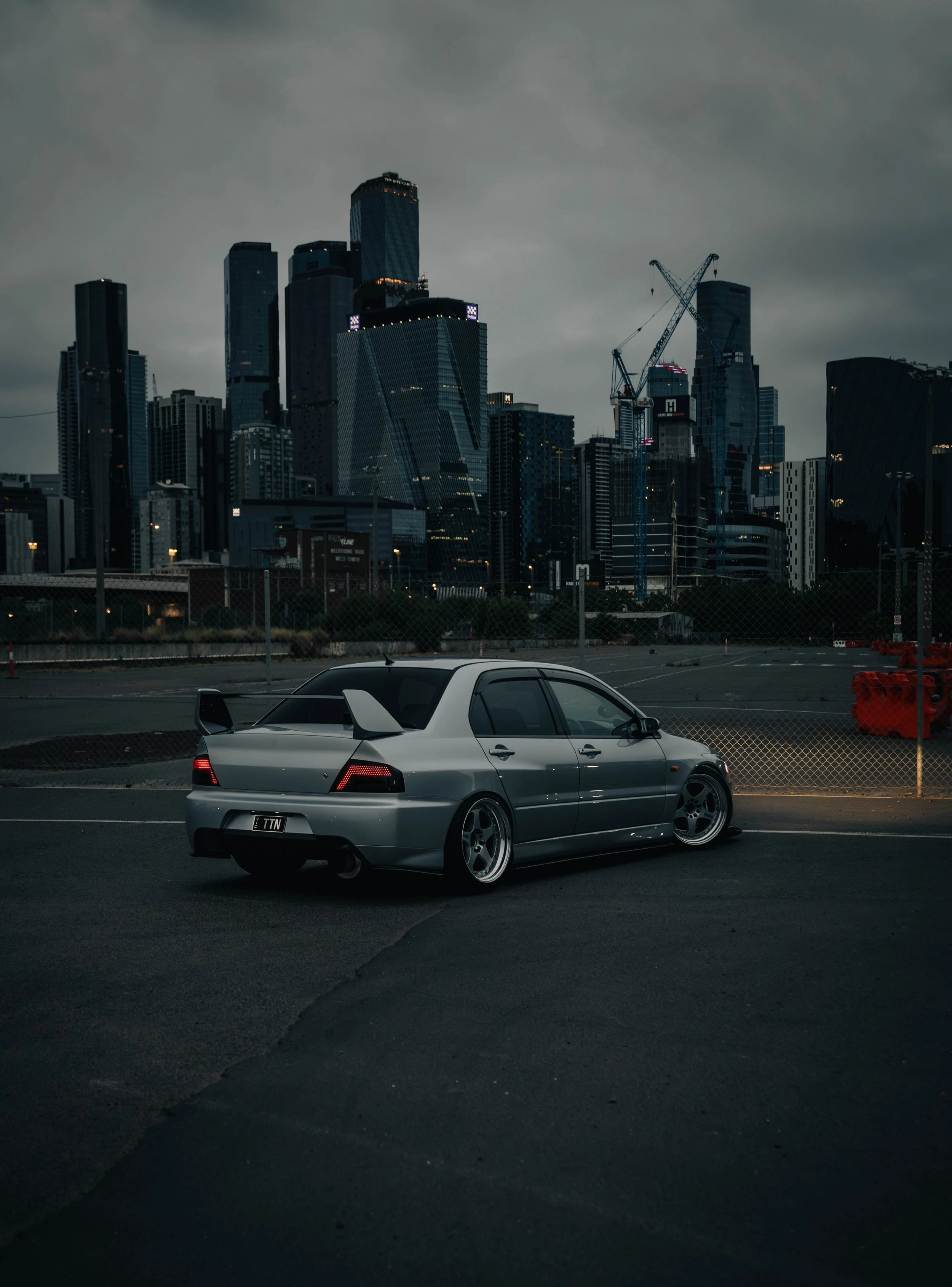 A silver sports car is parked in an empty parking lot with high-rise buildings and construction cranes in the background under a cloudy sky.