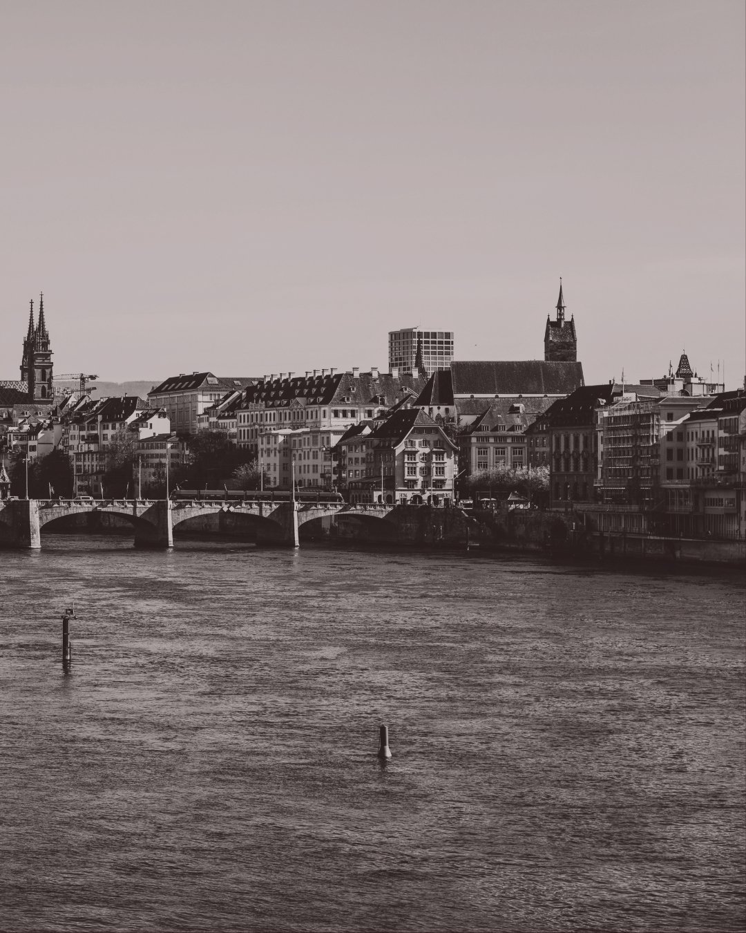 Ein Schwarz-Weiß-Foto einer Stadtansicht mit Fluss im Vordergrund, einer Brücke und historischen Gebäuden mit Türmen im Hintergrund.