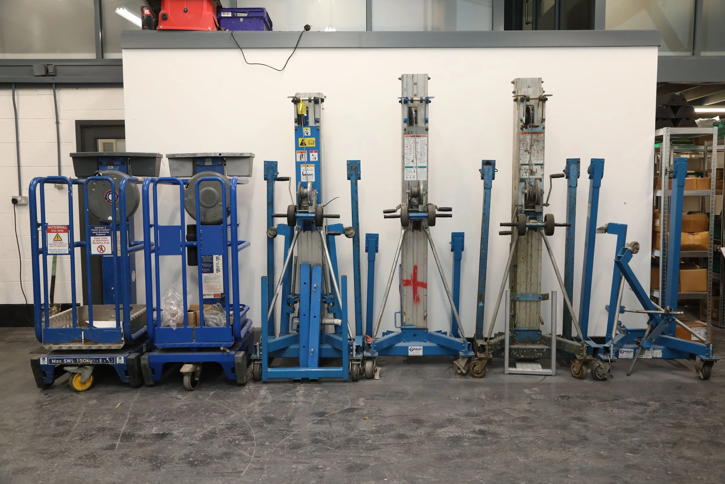 Row of various blue and gray mobile lifts and equipment carts on a concrete floor against a white wall.