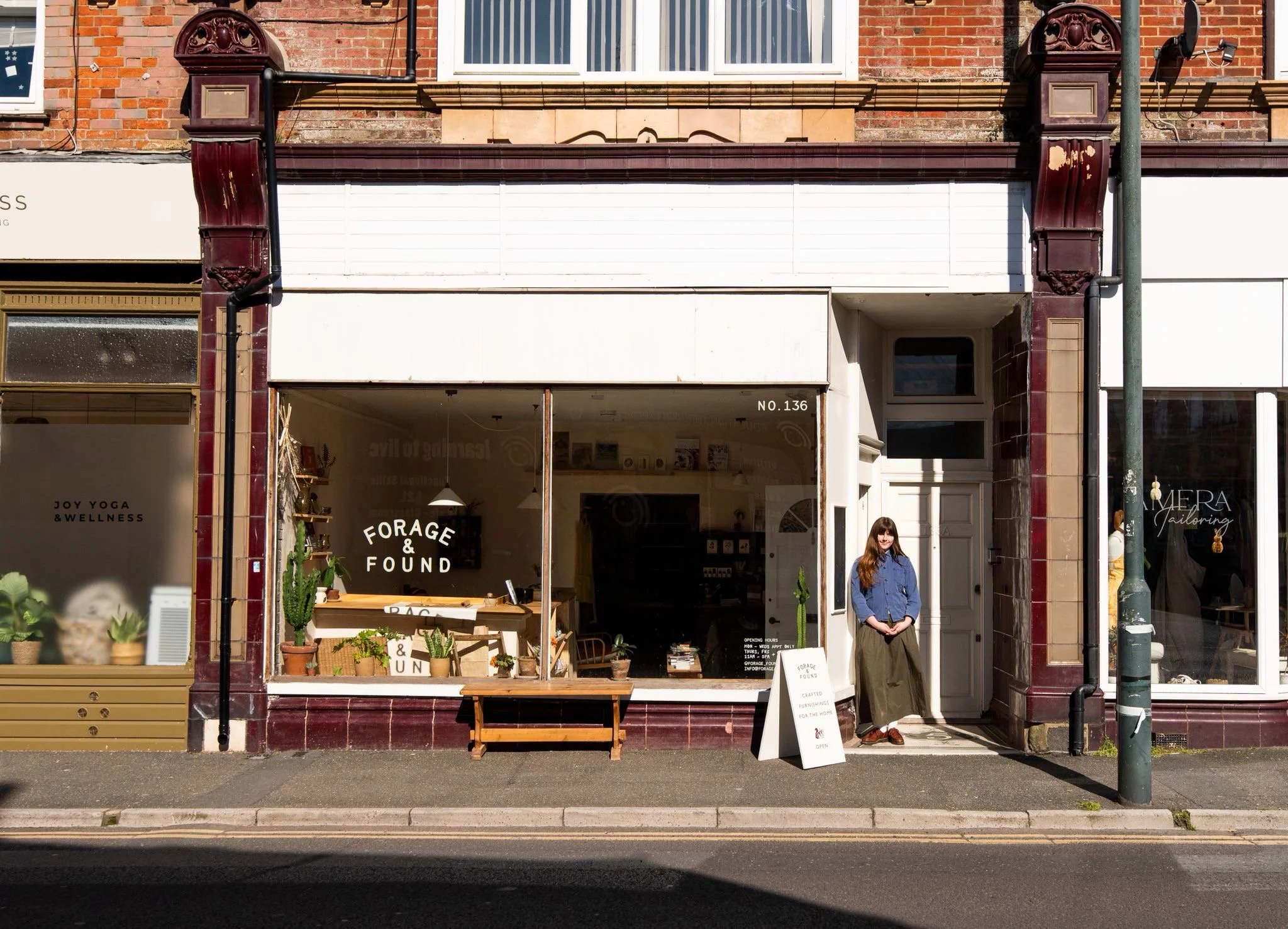A storefront with the sign 'Forage & Found' on the window, featuring potted plants and a wooden bench outside. A woman with long brown hair, wearing a blue top and long olive-green skirt, stands near the entrance. The adjacent window displays the sign 'Joy Yoga & Wellness.'
