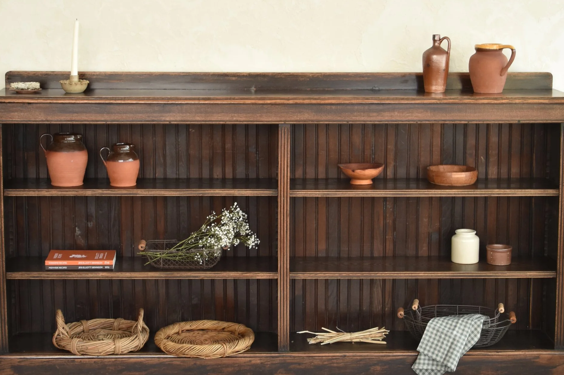 victorian Wooden panelled shelf with various pottery, bowls, books, a wicker basket, flowers, and linen tea towels in baskets.