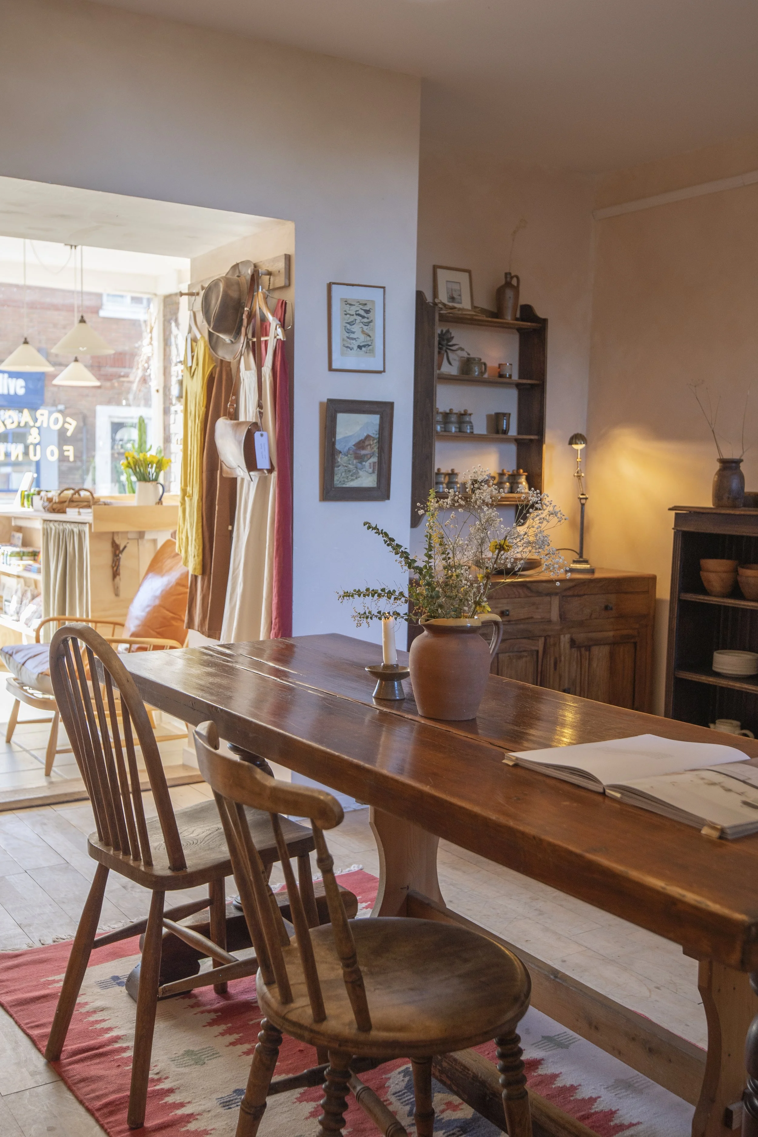 Interior of a cozy home with a wooden dining table, chairs, a vase with flowers, and warm lighting; a cozy corner with a dresser, shelves, and decorative items.