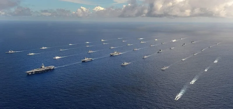 A fleet of ships, including aircraft carriers and smaller vessels, sailing in formation on the ocean.