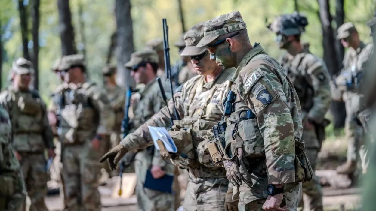 Group of soldiers in camouflage uniforms and tactical gear standing outdoors in a wooded area, some wearing sunglasses and hats, engaging in a discussion.