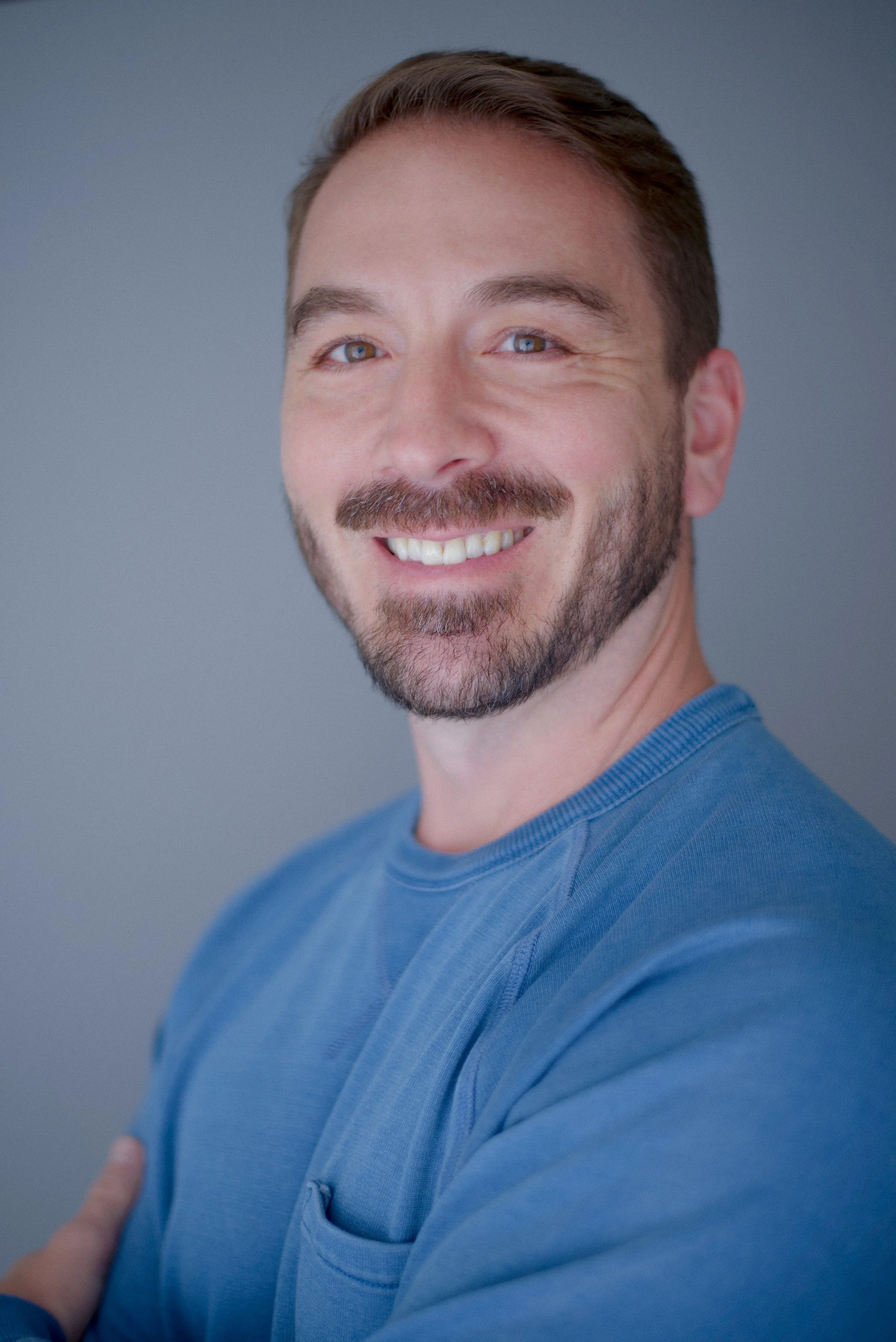 Justin Riestra, an actor and filmmaker based in Santa Monica, California, looks toward the camera, smiling. He's wearing a light blue sweater, he has short brown hair, brown eyes, and a well trimmed beard and mustache.