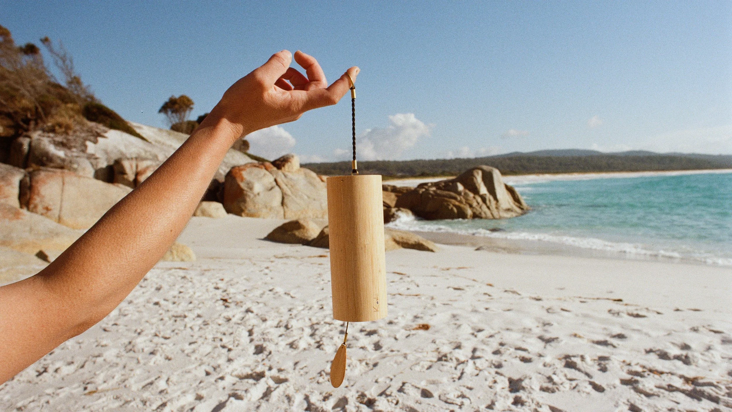 A person holding a wooden wind chime with a seaside background, rocks, and blue sky.